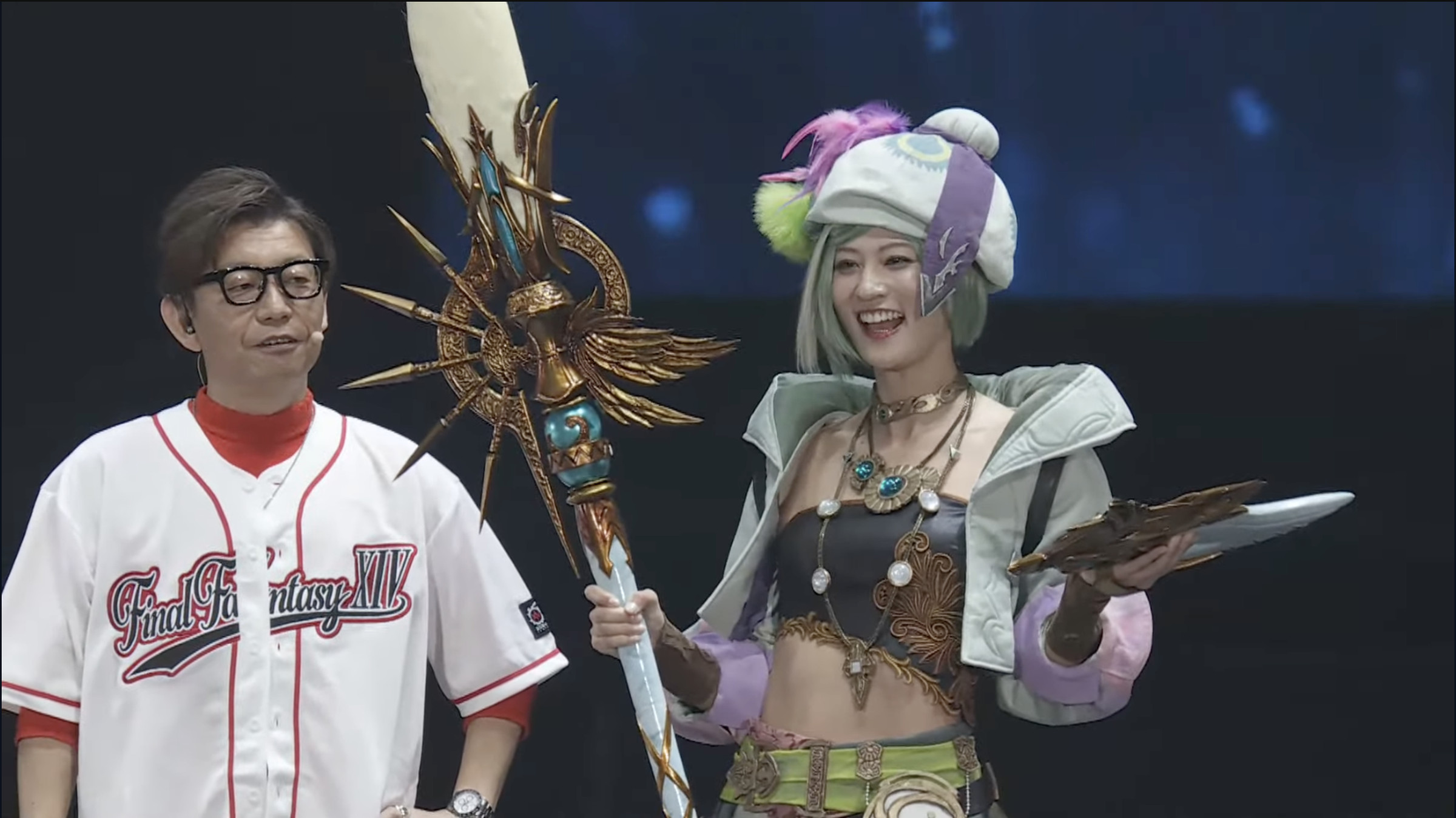 Screenshot from the Final Fantasy XIV Fan Fest event featuring FFXIV game producer Naoki Yoshida on the left wearing a Final Fantasy XIV red and white baseball jersey standing next to a model cosplaying the pictomancer job class.