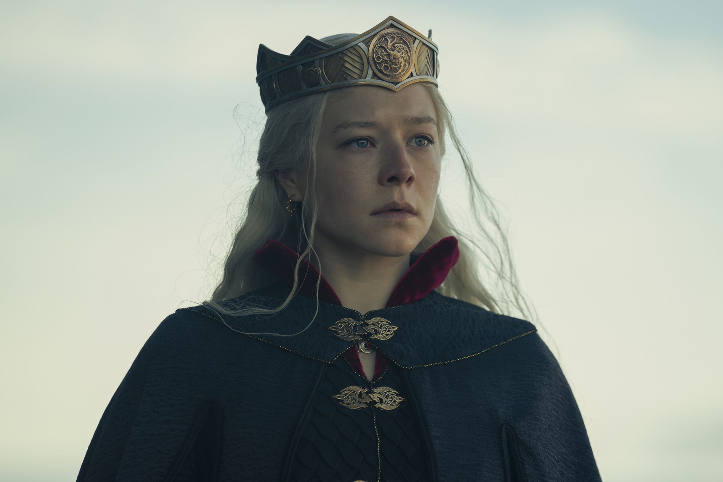 A platinum blonde wearing a navy blue ceremonial cloak, a crown, and a deadened facial expression.