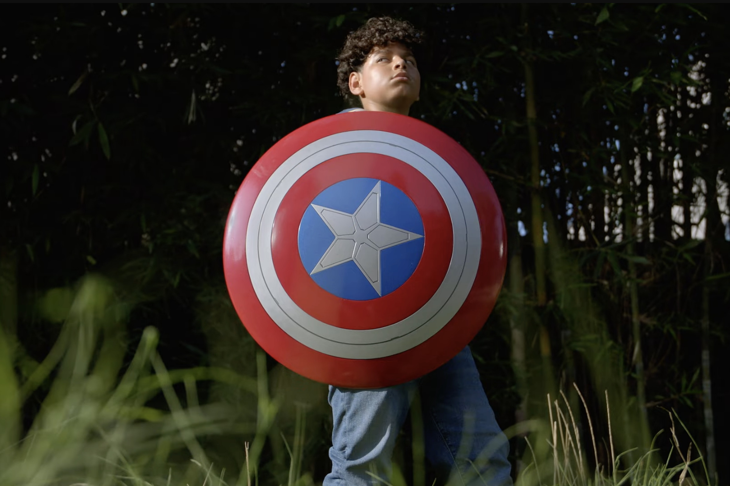 Image of young boy outside standing in grass looking heroically into the distance while holding Captain America’s sheild