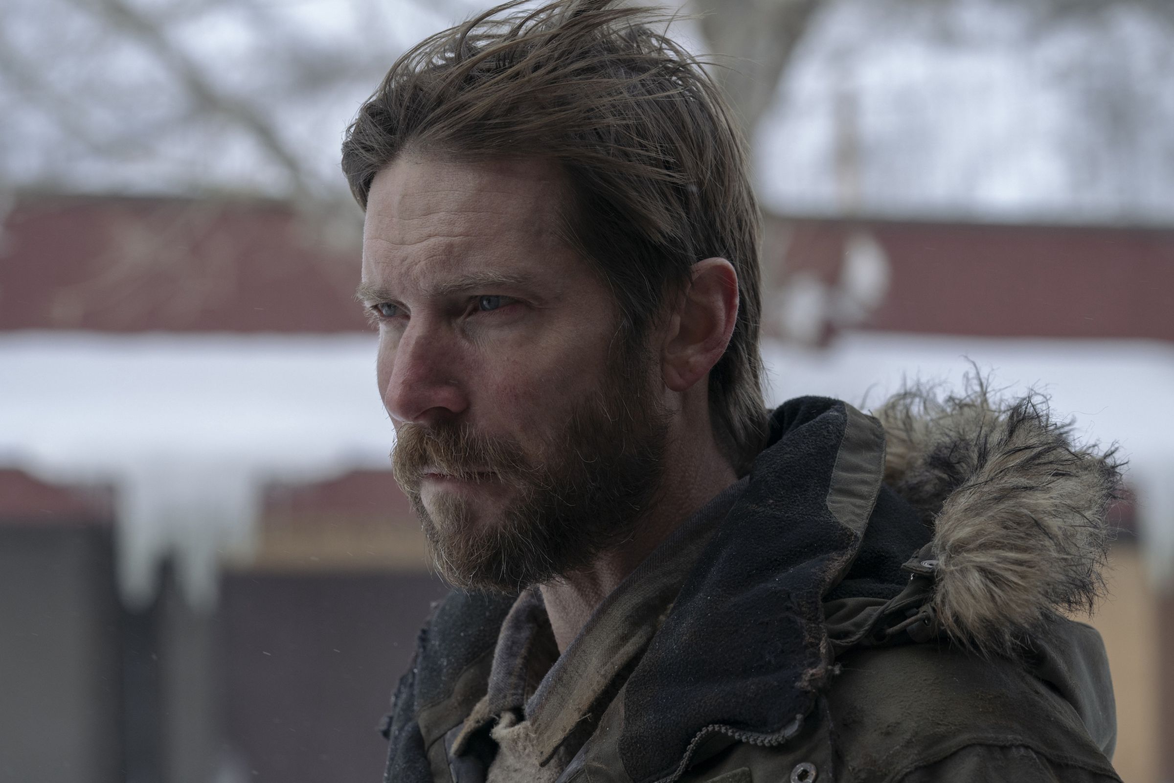 A bearded man with brown hair wearing a winter coat, and standing outside in the cold.