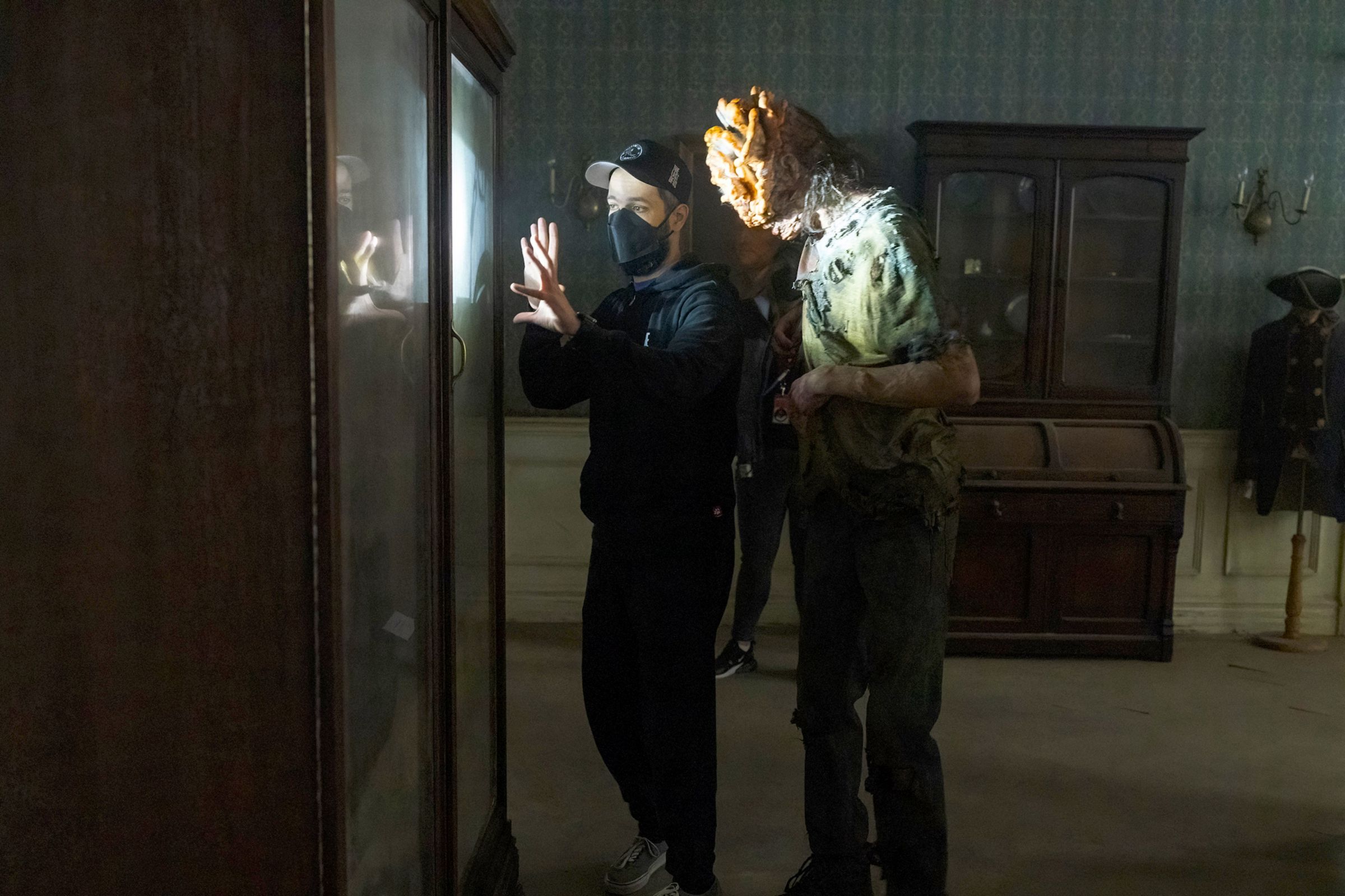 Two men on a film set standing in front of a display case. Closest to the case is a man in a black hoodie, jeans, a baseball cap, and a black mask as he gestures towards something while giving the other man. The second man, standing just behind the first is wearing a mottled pair of pants, a shirt, and a face full of prosthetics to make it look like his head has been colonized by an aggressive fungal growth.