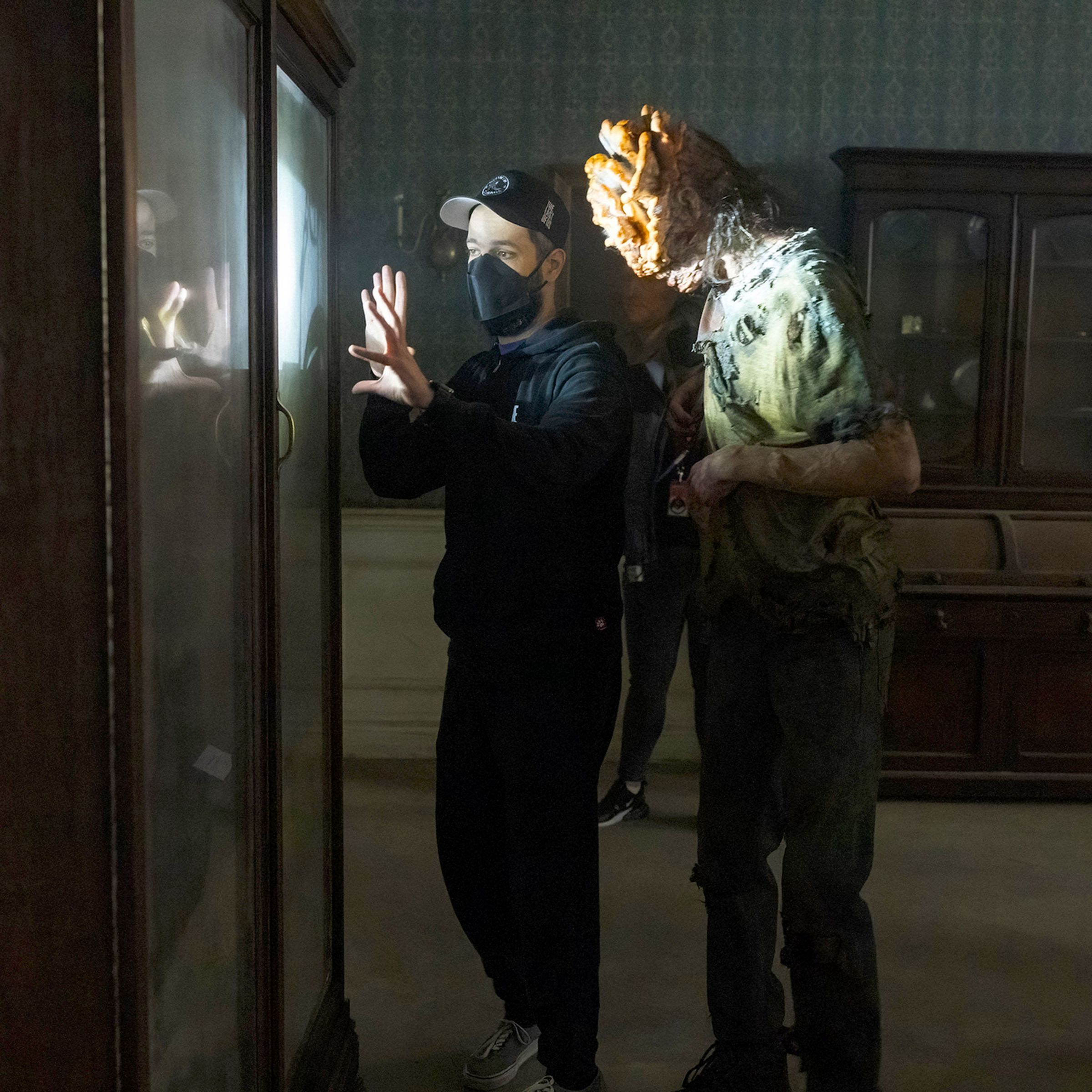 Two men on a film set standing in front of a display case. Closest to the case is a man in a black hoodie, jeans, a baseball cap, and a black mask as he gestures towards something while giving the other man. The second man, standing just behind the first is wearing a mottled pair of pants, a shirt, and a face full of prosthetics to make it look like his head has been colonized by an aggressive fungal growth.