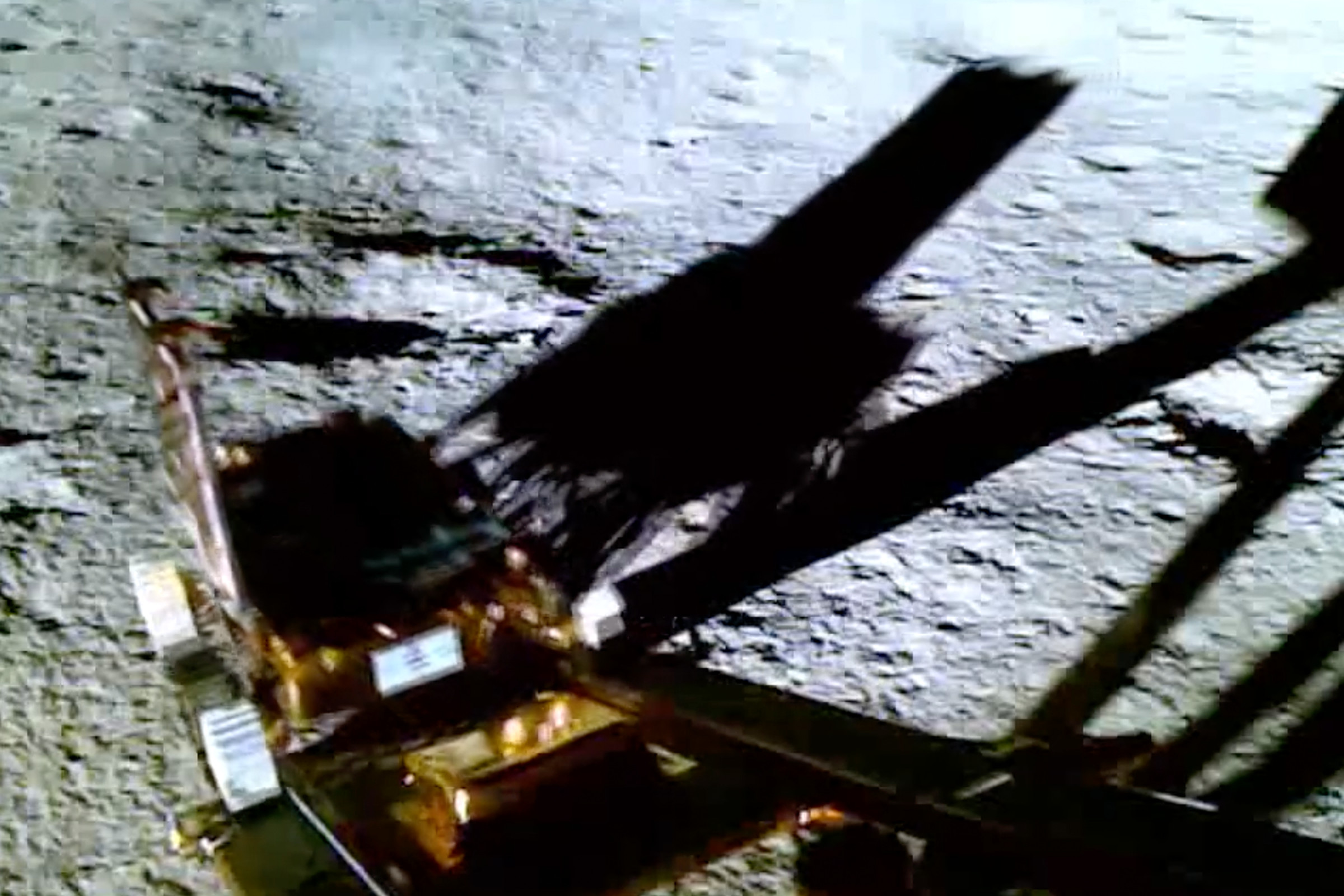 An image of the Chandrayaan-3 rover as it reaches the end of the landing ramp. In the background, the pocked surface of the Moon fills about the top two-thirds of the image. The picture is in color, showing the copper-colored foil that covers the sides of the rover.