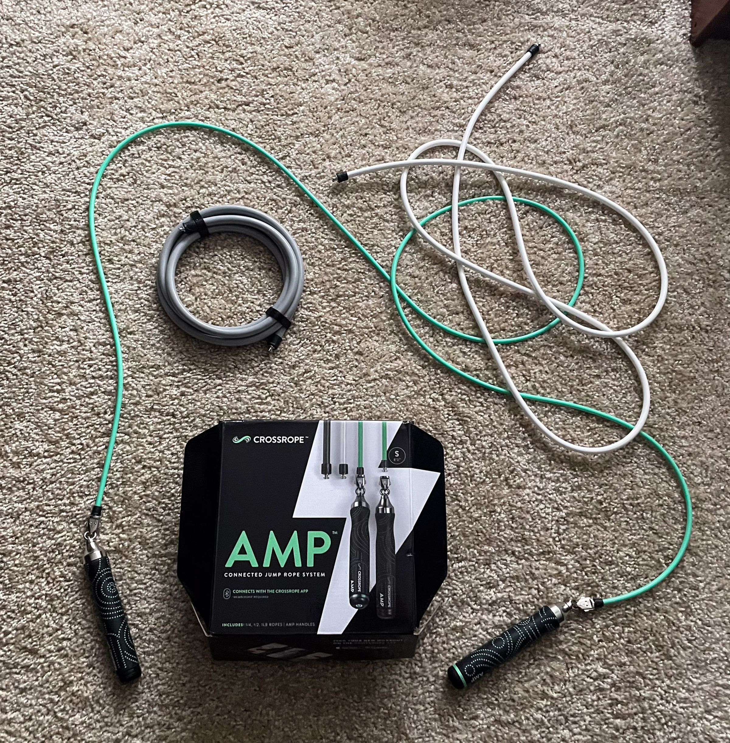 The Crossrope AMP Jump Rope set box surrounded by its three green, gray, and white weighted jump ropes, with the AMP handled attached to the green one.