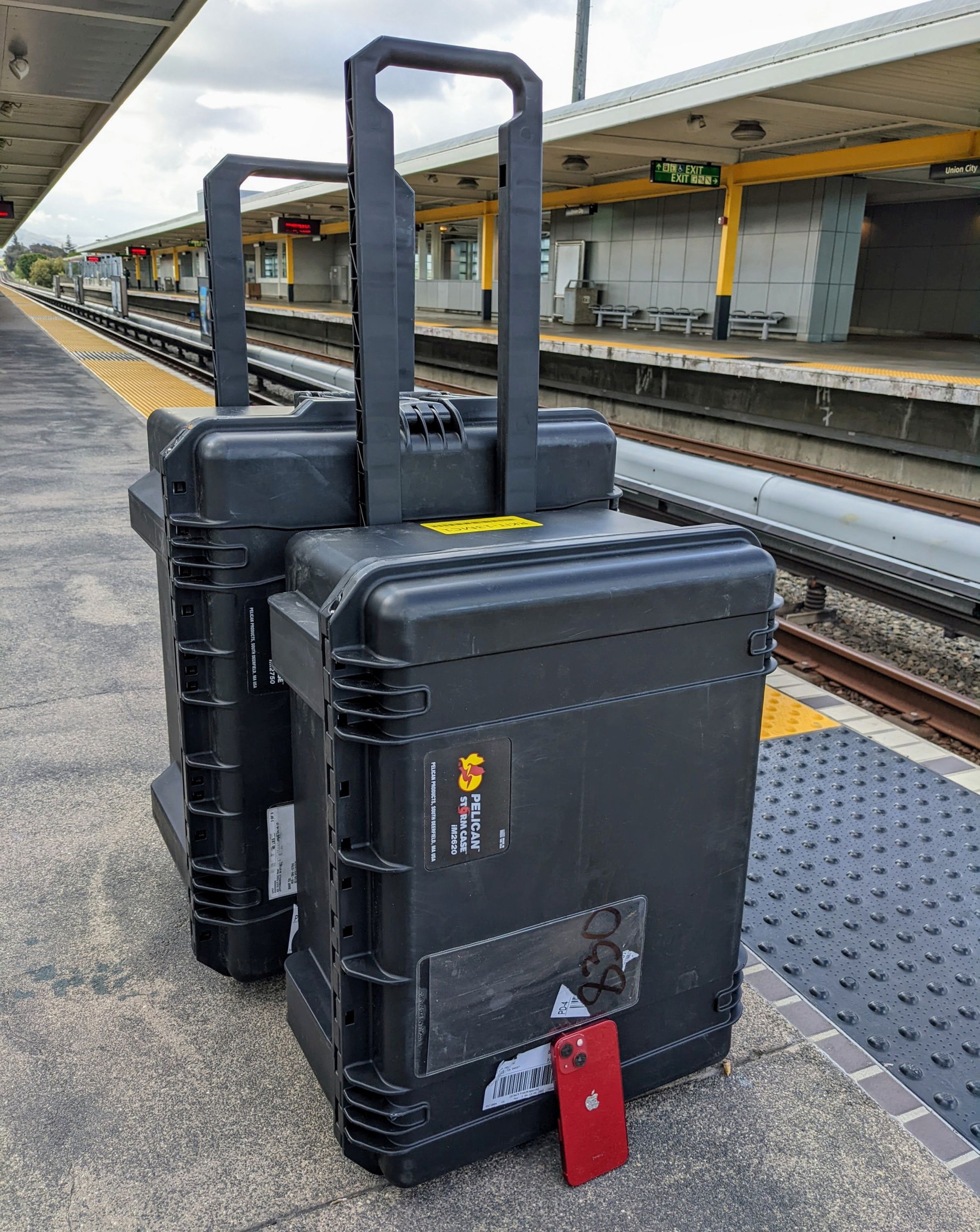 Two Pelican cases sit on a train platform, with a tiny red iPhone 13 mini next to them for scale.