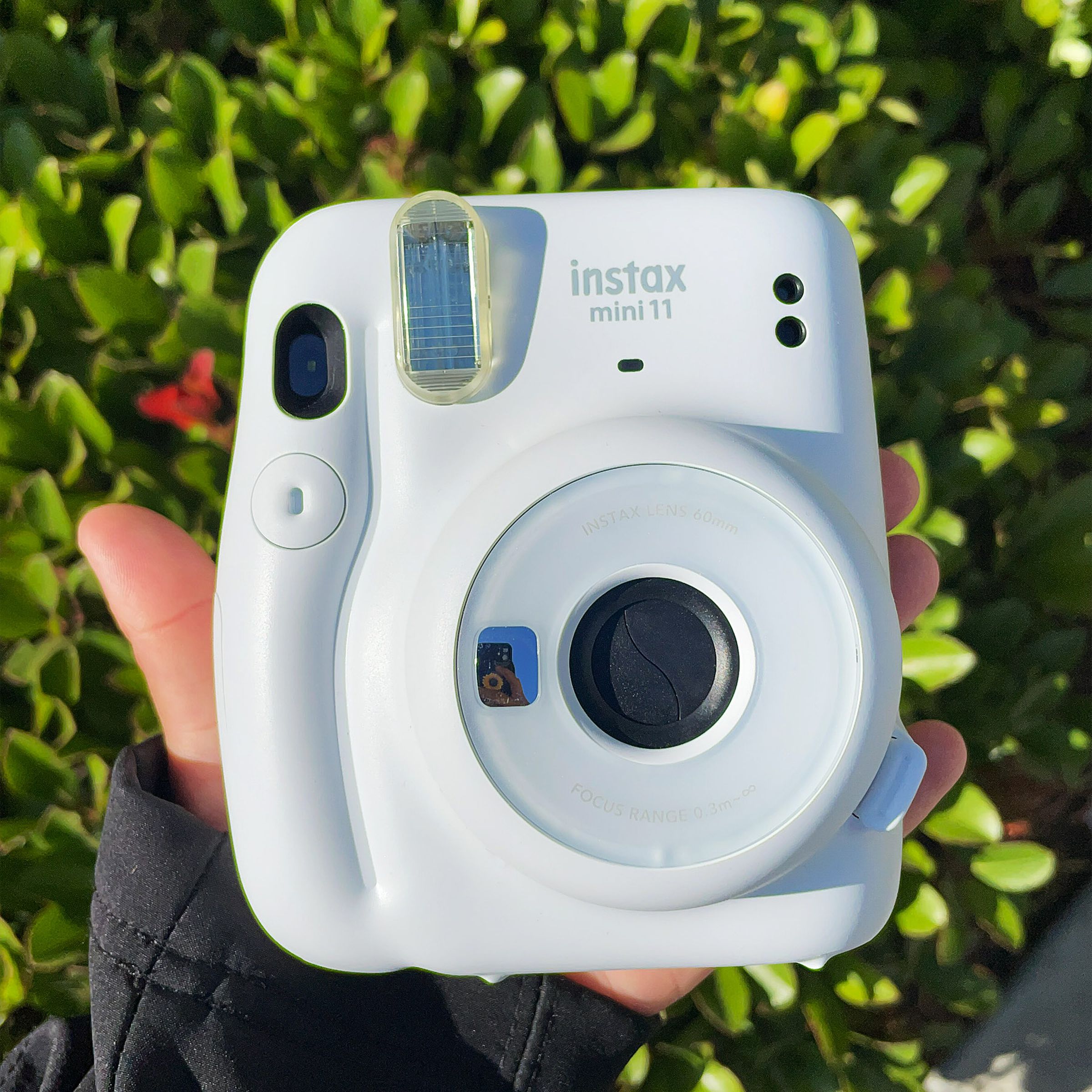 A hand holding Fujifilm’s white Instax Mini 11 instant camera in front of a bush outside.
