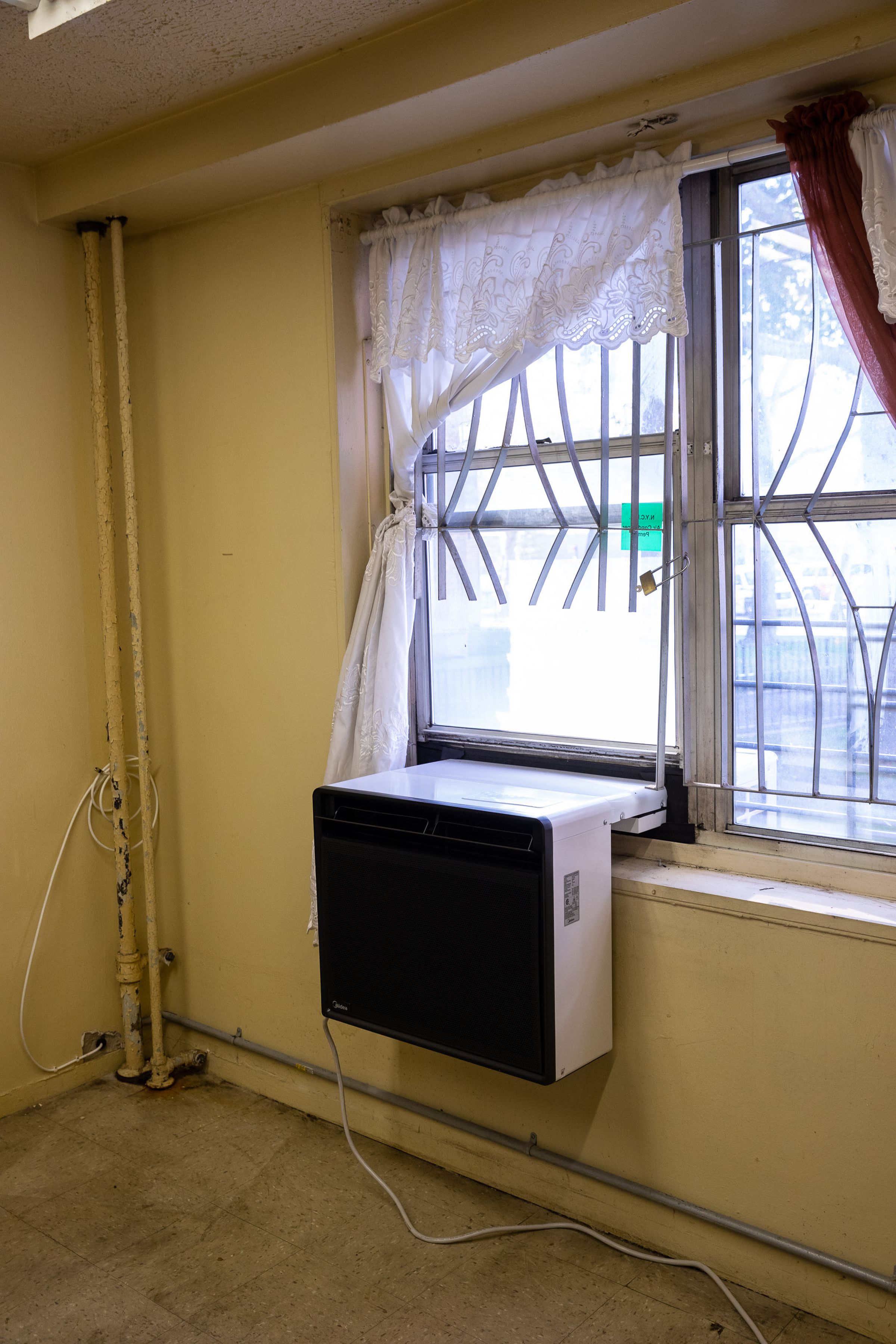 A rectangular heat pump sits in a window sill with off-white lace curtains tucked behind it.