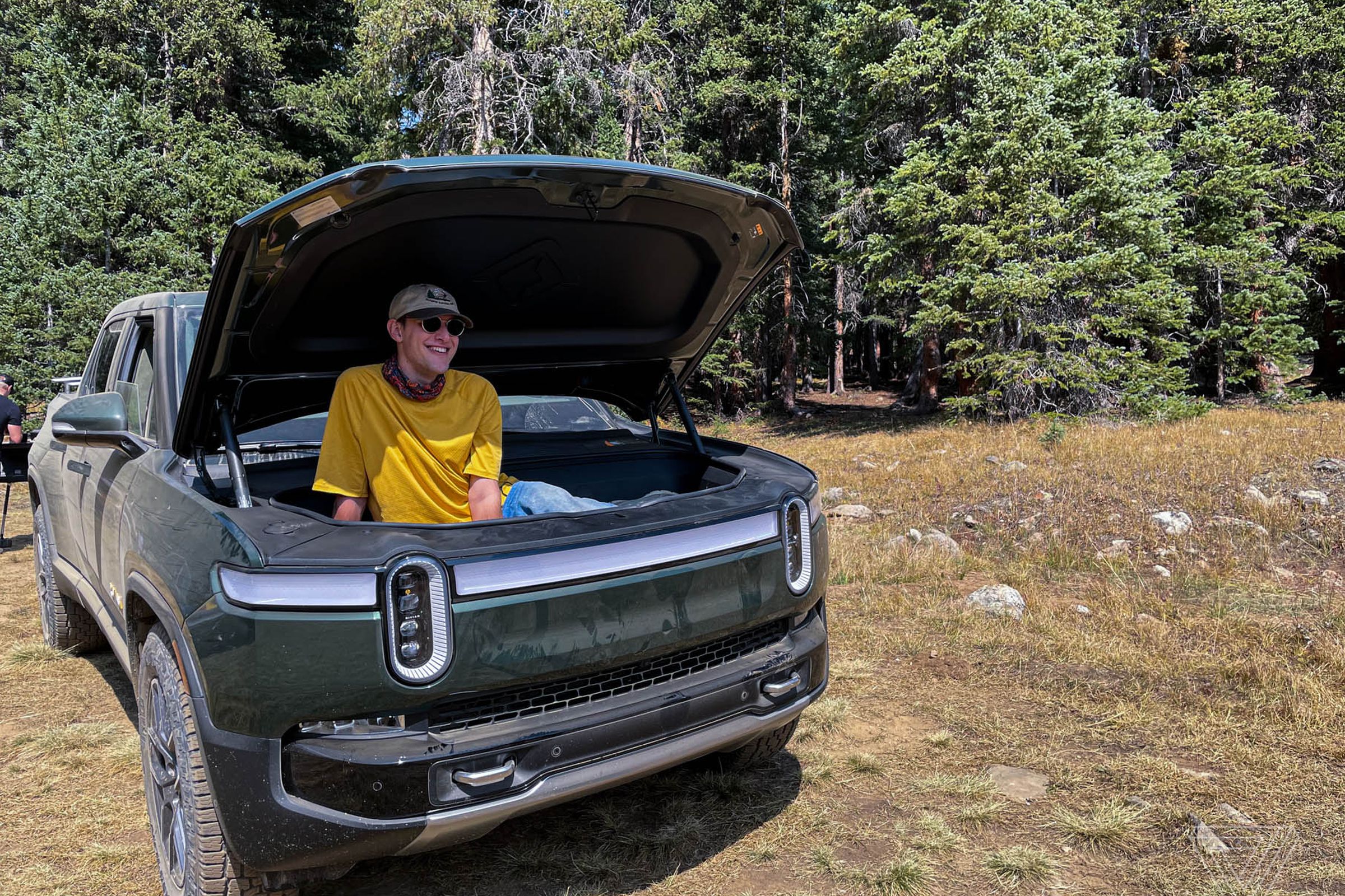 The Verge’s Mitchell Clark sits inside the Rivian R1T’s front trunk or frunk with the hood open as he grins at a trail he’ll embark on very soon.