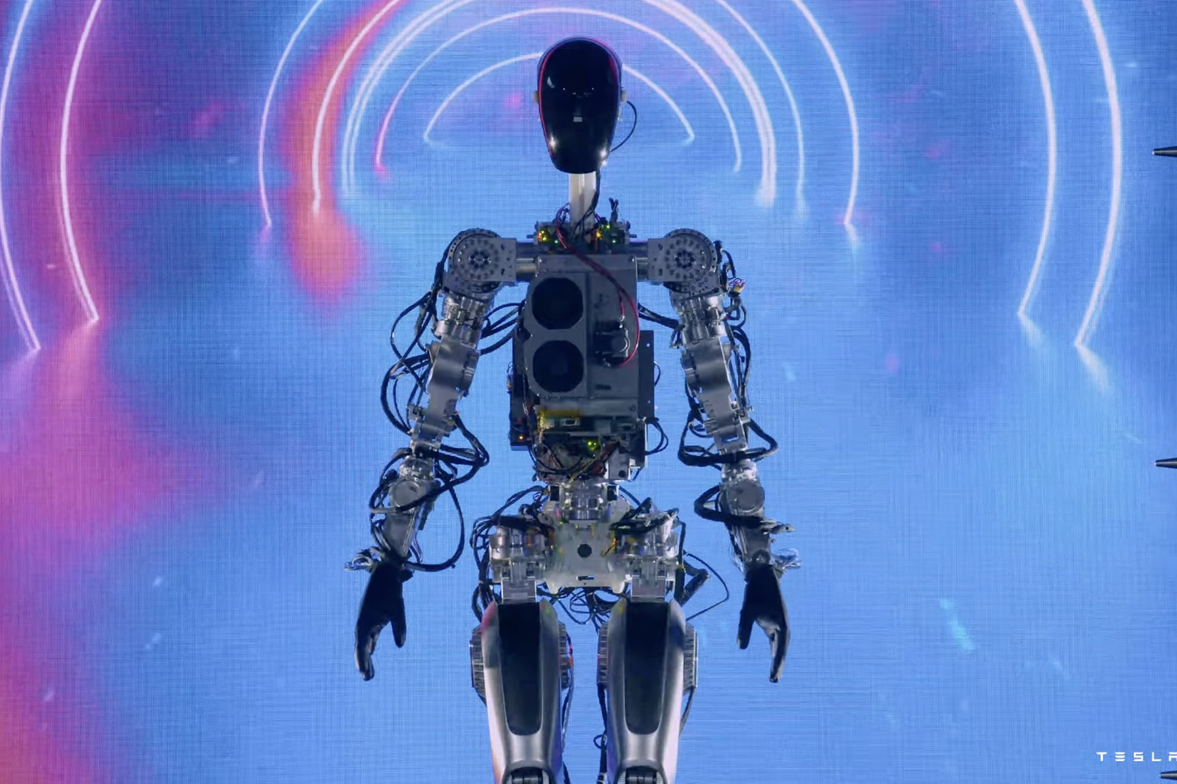A photograph of a humanoid robot with exposed wiring and innards stands in front of a colorful screen.  