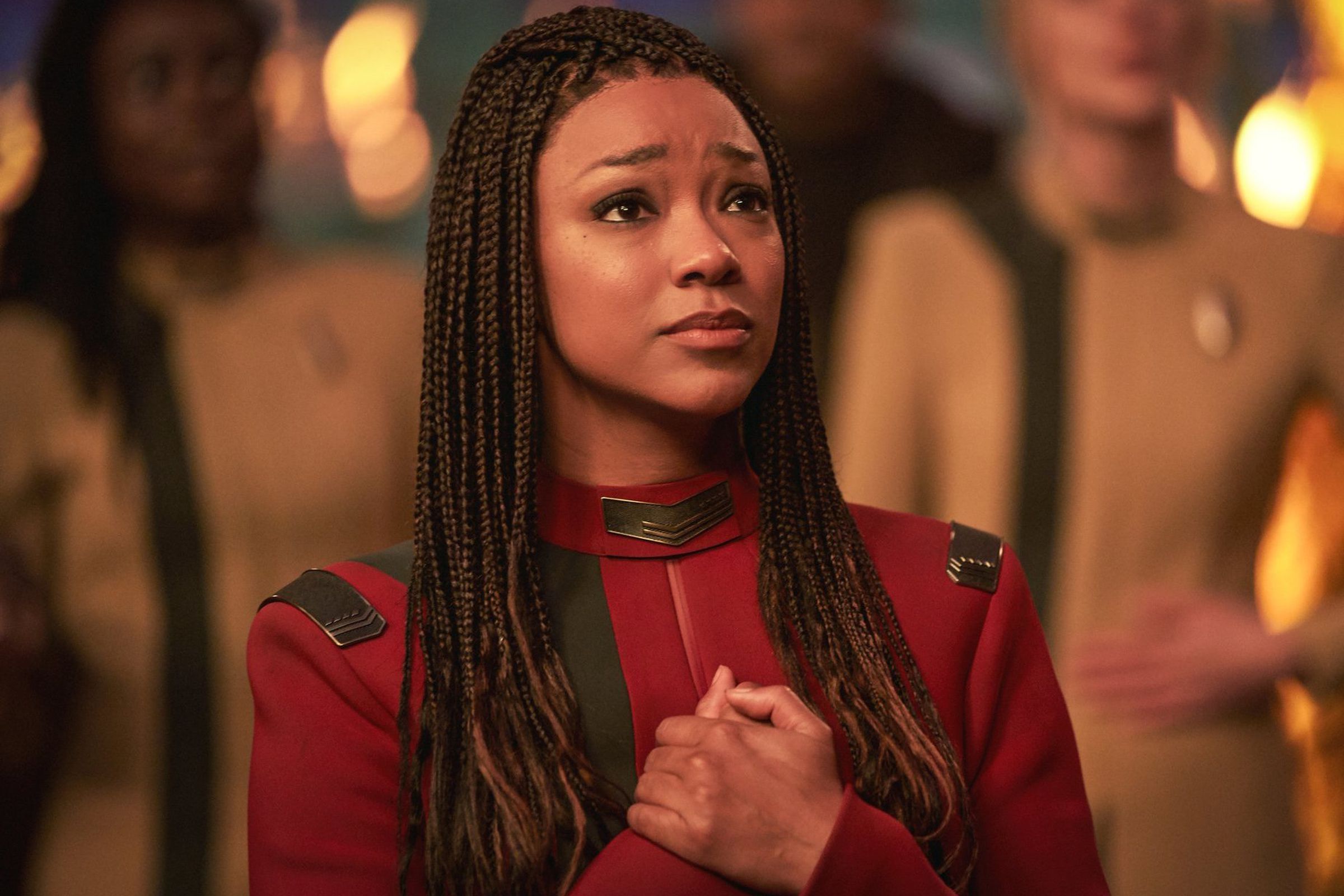 A Black woman with long braids wearing a red uniform and clasping her hands together in front of her chest.