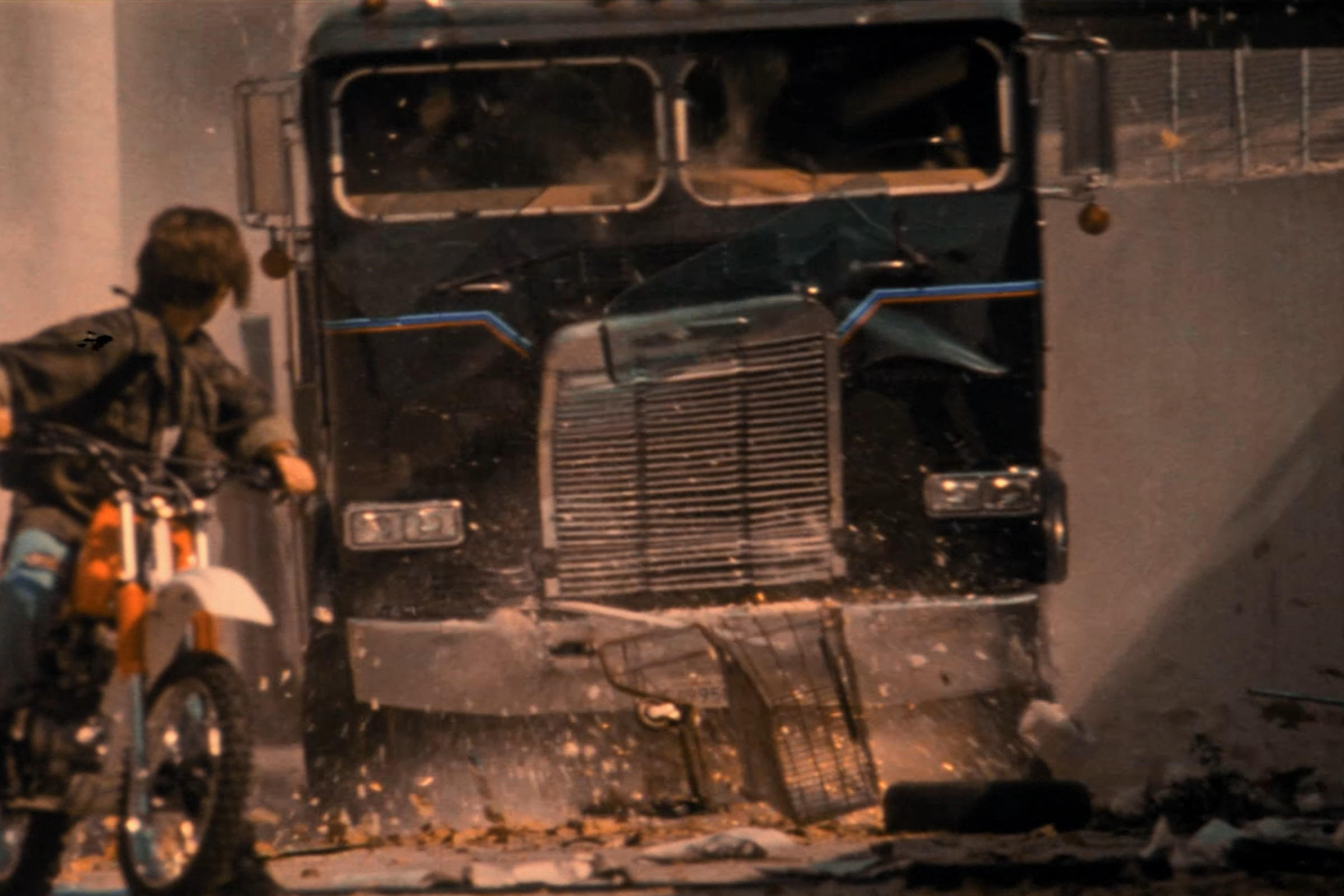 A screenshot from Terminator 2 showing a person on a dirtbike in the foreground and a flat-nosed semi truck in the background, approaching him.