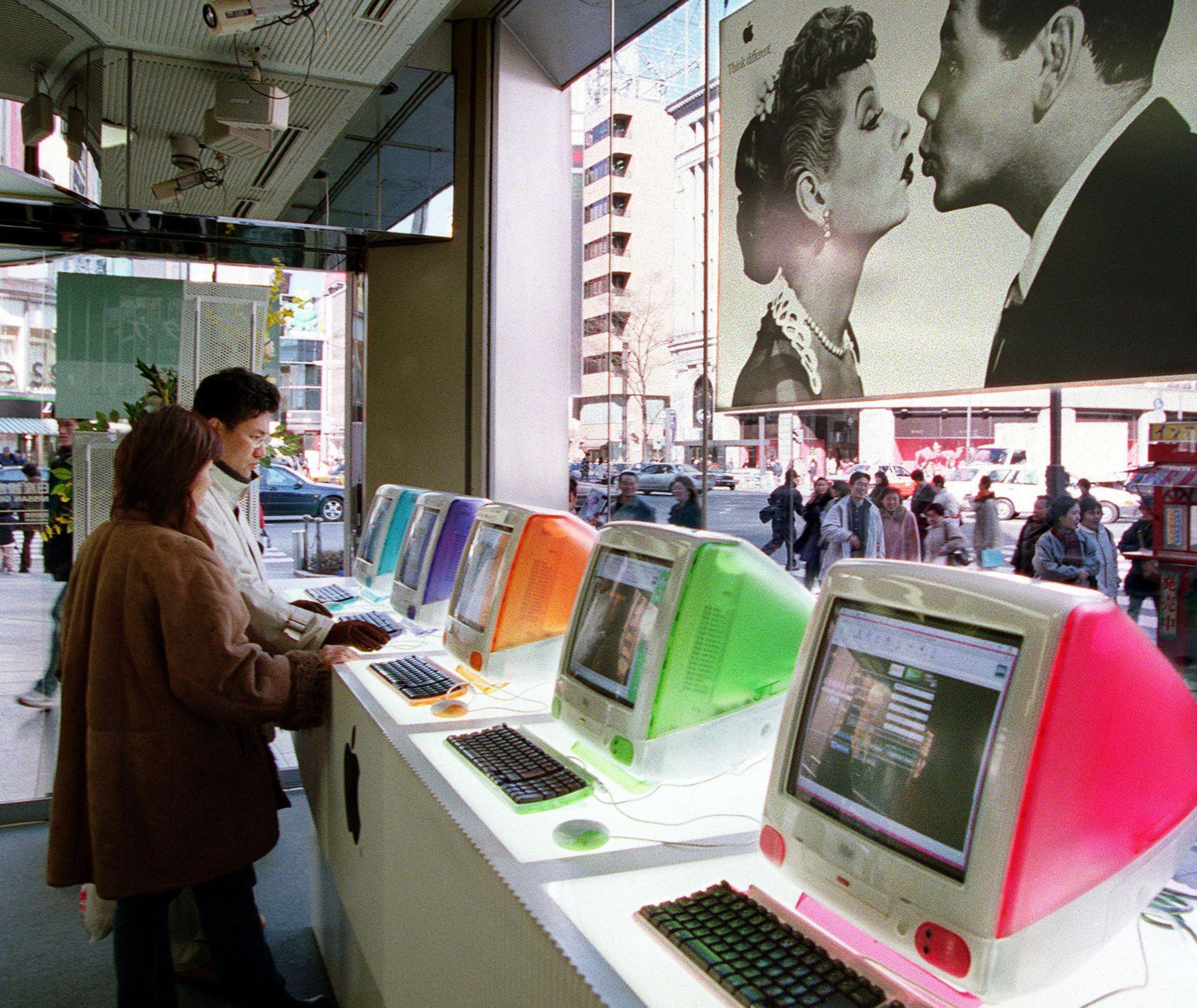 Several iMacs sit on a counter in a showroom as two people look at them.