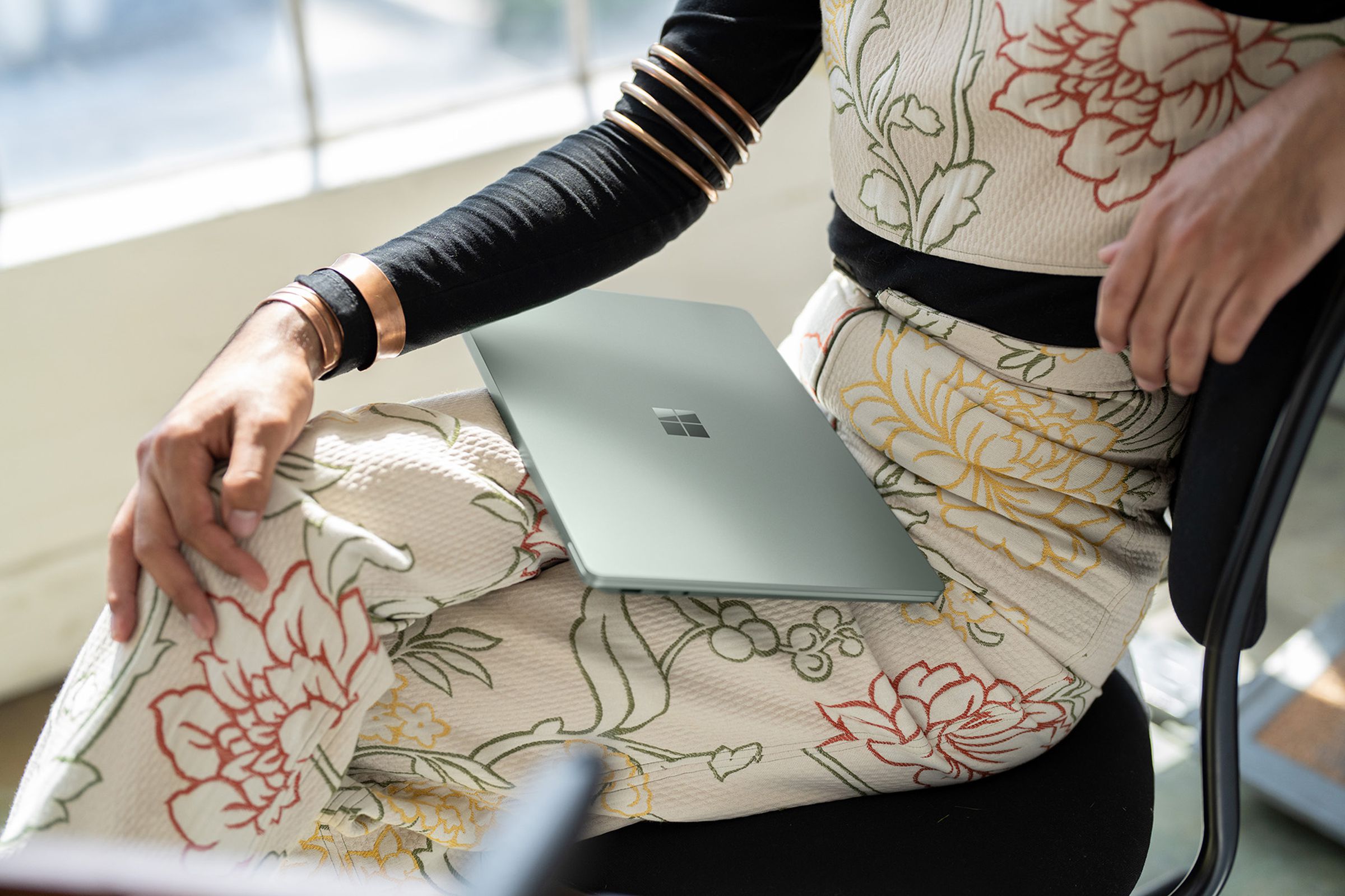A seated user in a floral bodysuit holds a closed Surface Laptop 5 in their lap with the Microsoft logo visible.