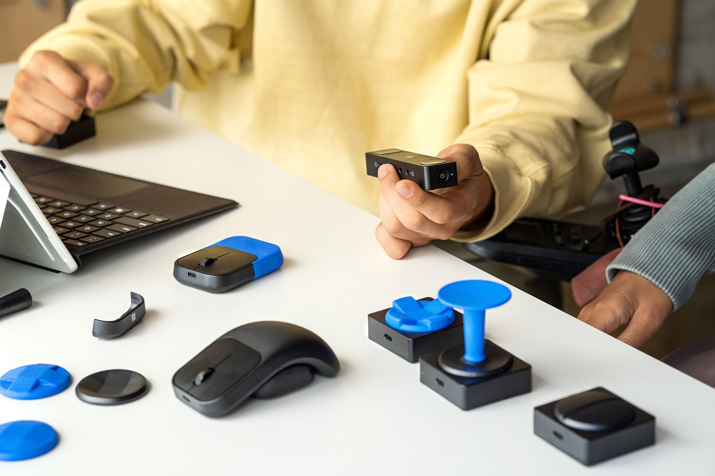 A selection of Microsoft Adaptive Accessories being used by two people who are sat at a desk, using a laptop.