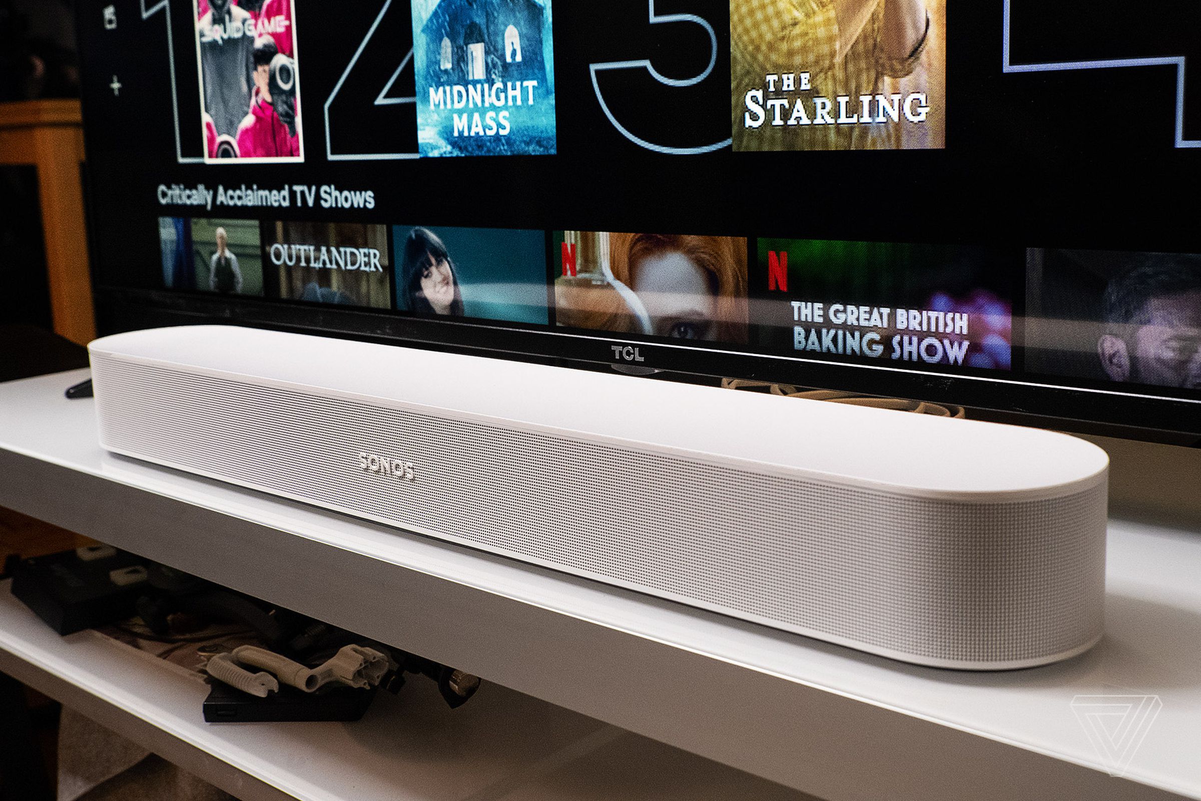 The white-colored, second-generation Sonos Beam sits on a white entertainment center. This is a close-up shot, showing its grille that lets out sound. The soundbar is sitting directly in front of a TCL flat-screen TV.