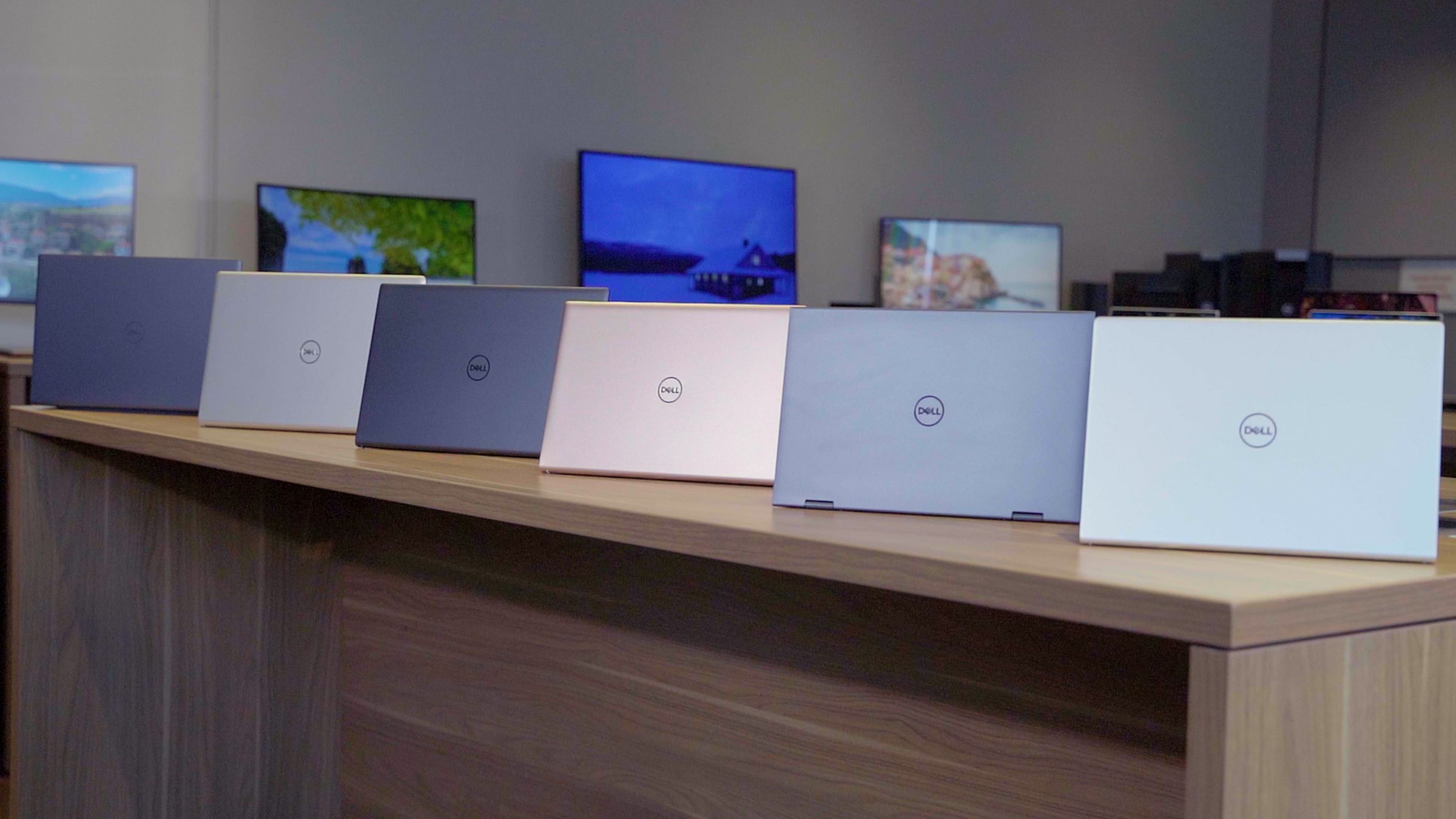 Six Dell Inspiron models sit in a line, open, with their lids facing the camera, on a table in a retail store.