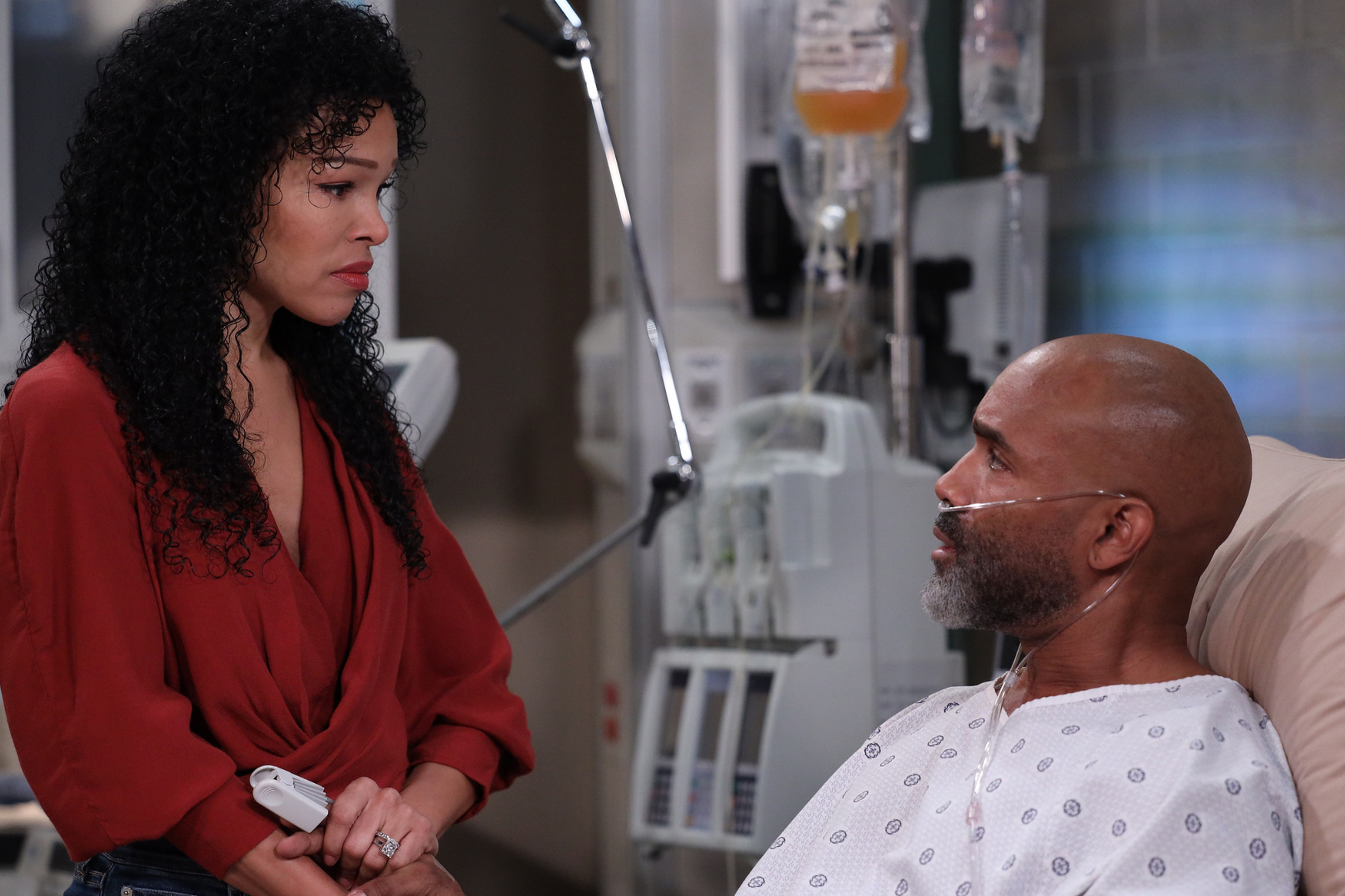 Photo from General Hospital – Episode “15264” featuring a brown woman with curly hair wearing a red shirt speaking to a bald African American man in a hospital bed wearing a hospital gown.