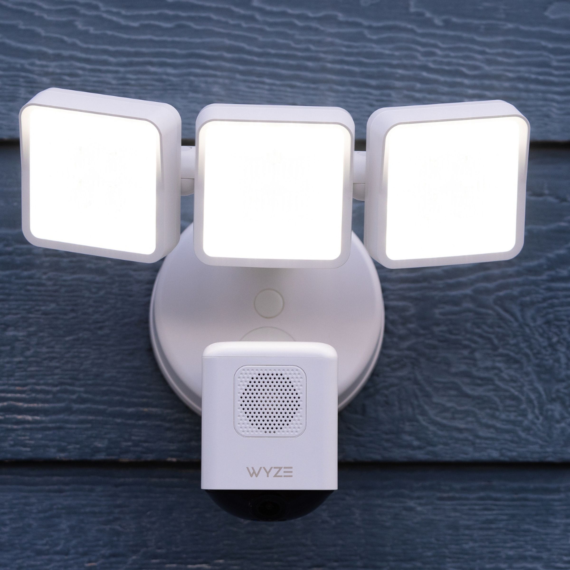 A photograph of the Wyze Cam Floodlight Pro in white mounted on a blue-gray wooden wall.