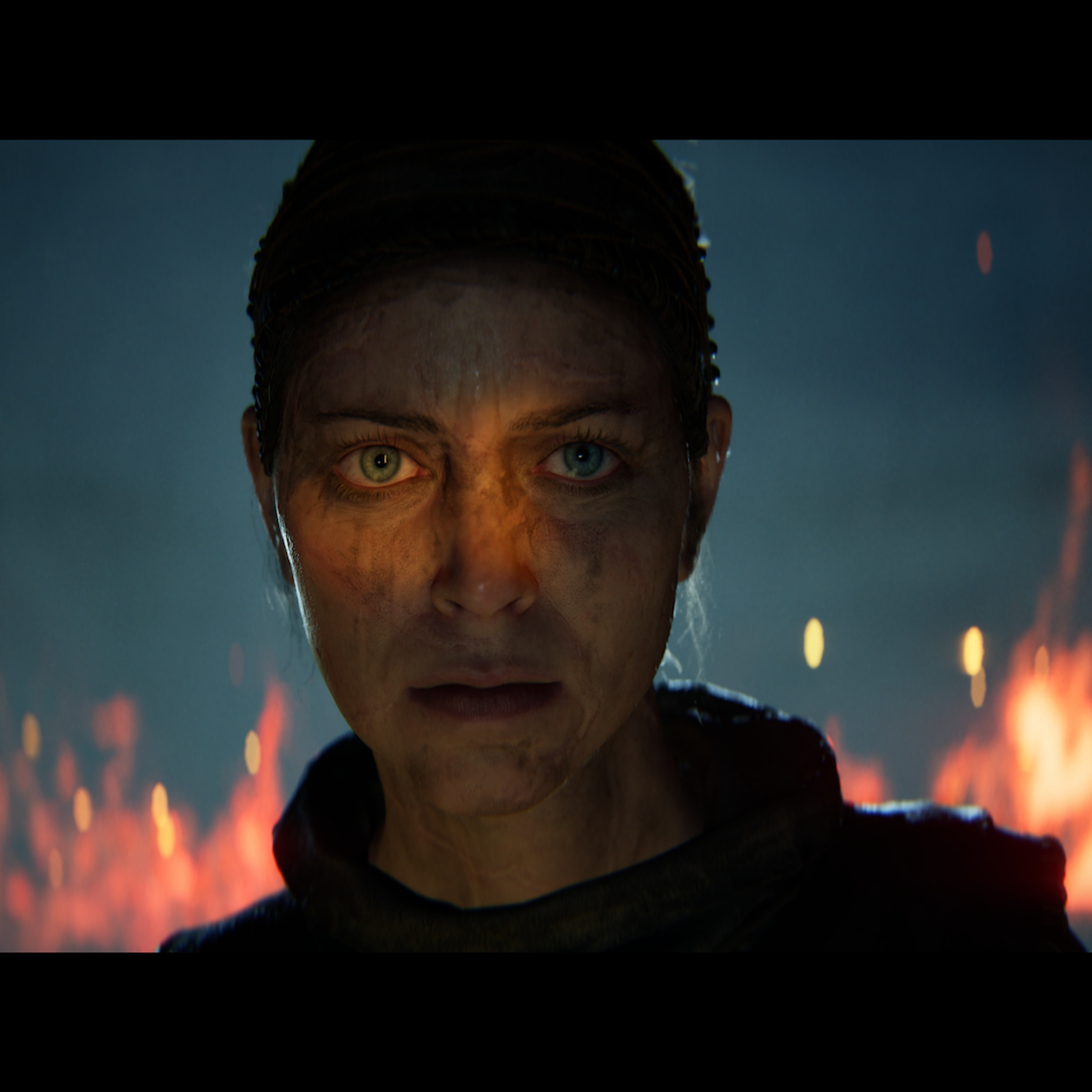 Screenshot from Senua’s Saga: Hellblade II featuring a close up shot of Senua, a woman covered in grime with flames burning behind her.