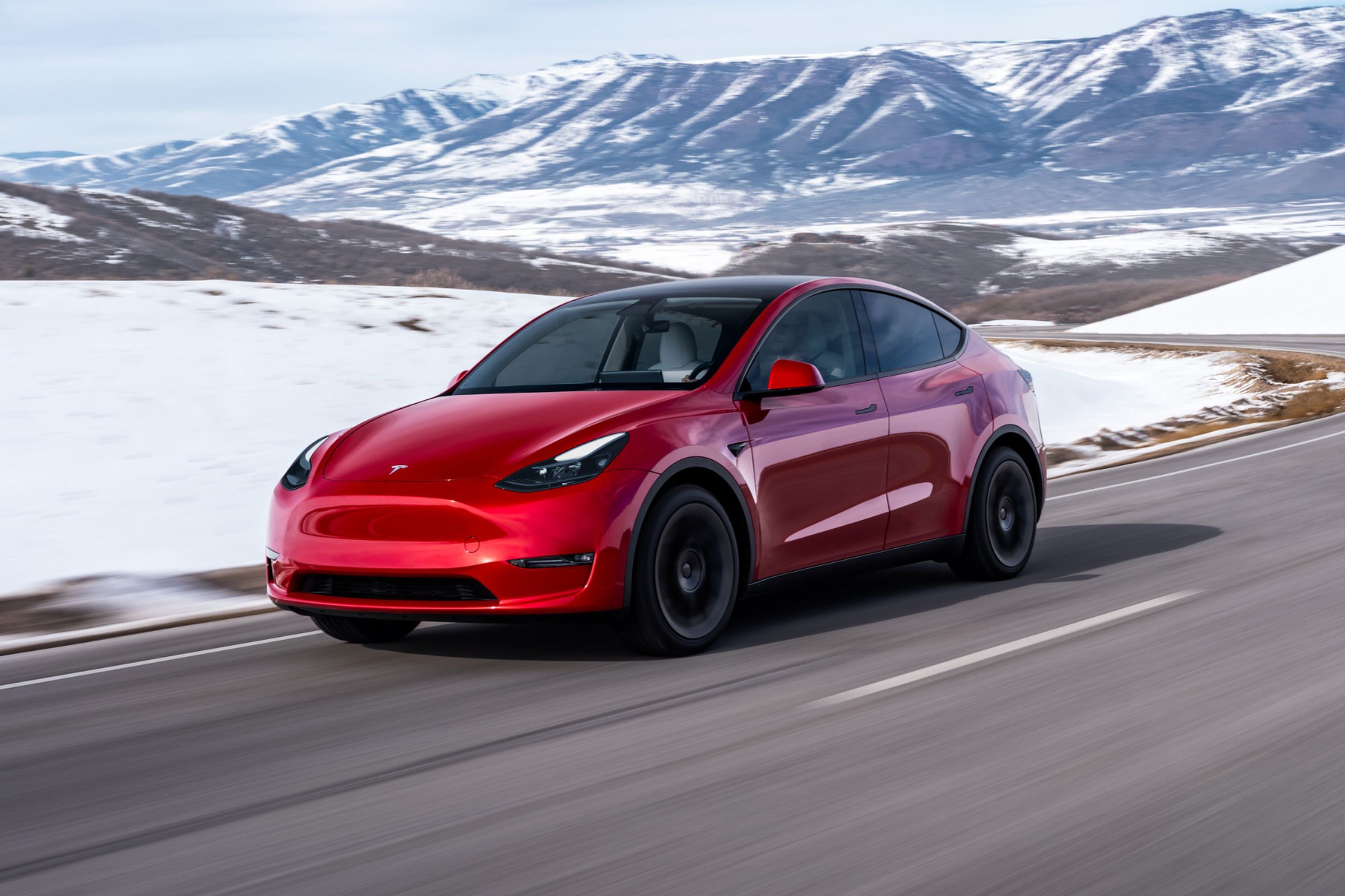 A red Tesla Model Y driving through a snowy, mountainous landscape.