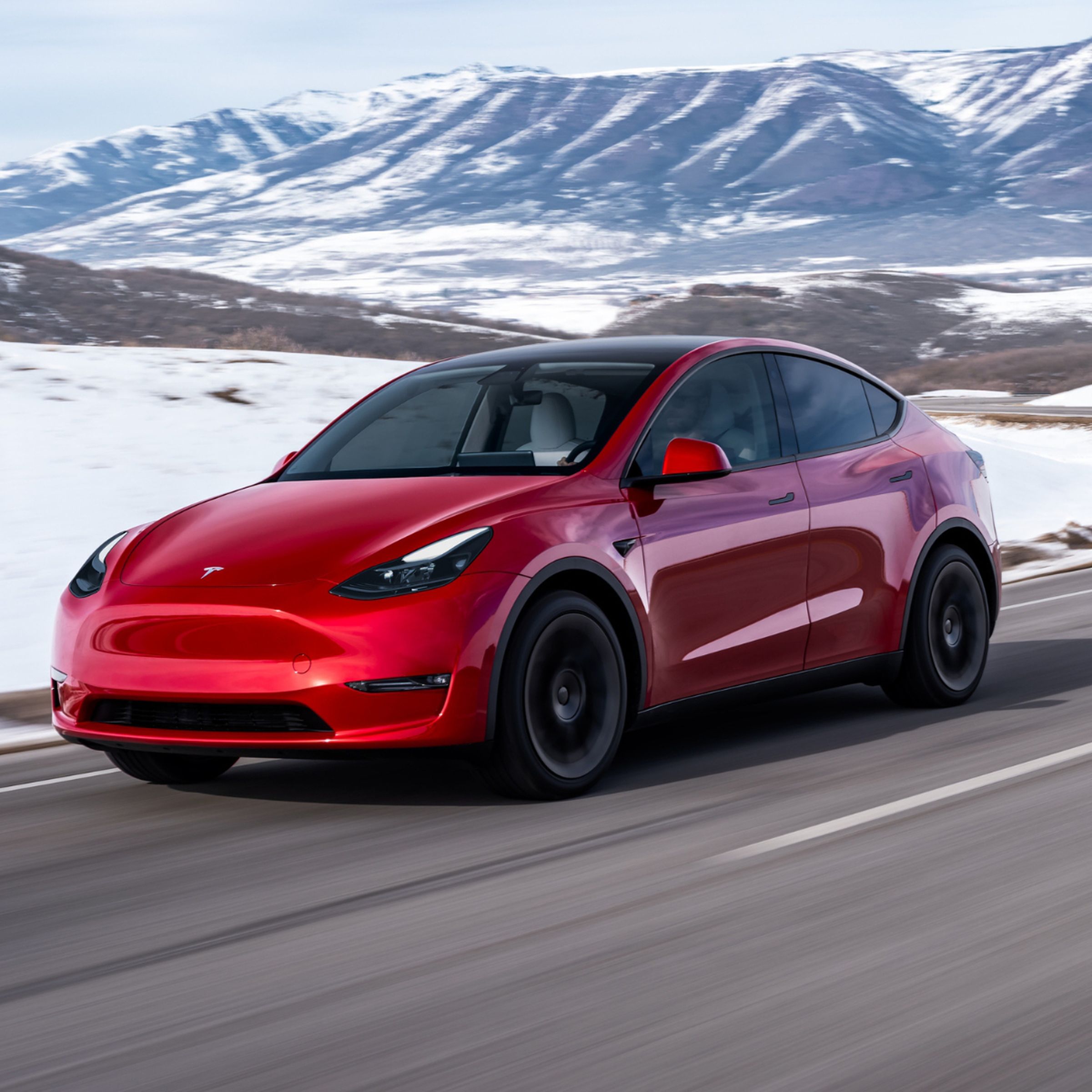 A red Tesla Model Y driving through a snowy, mountainous landscape.