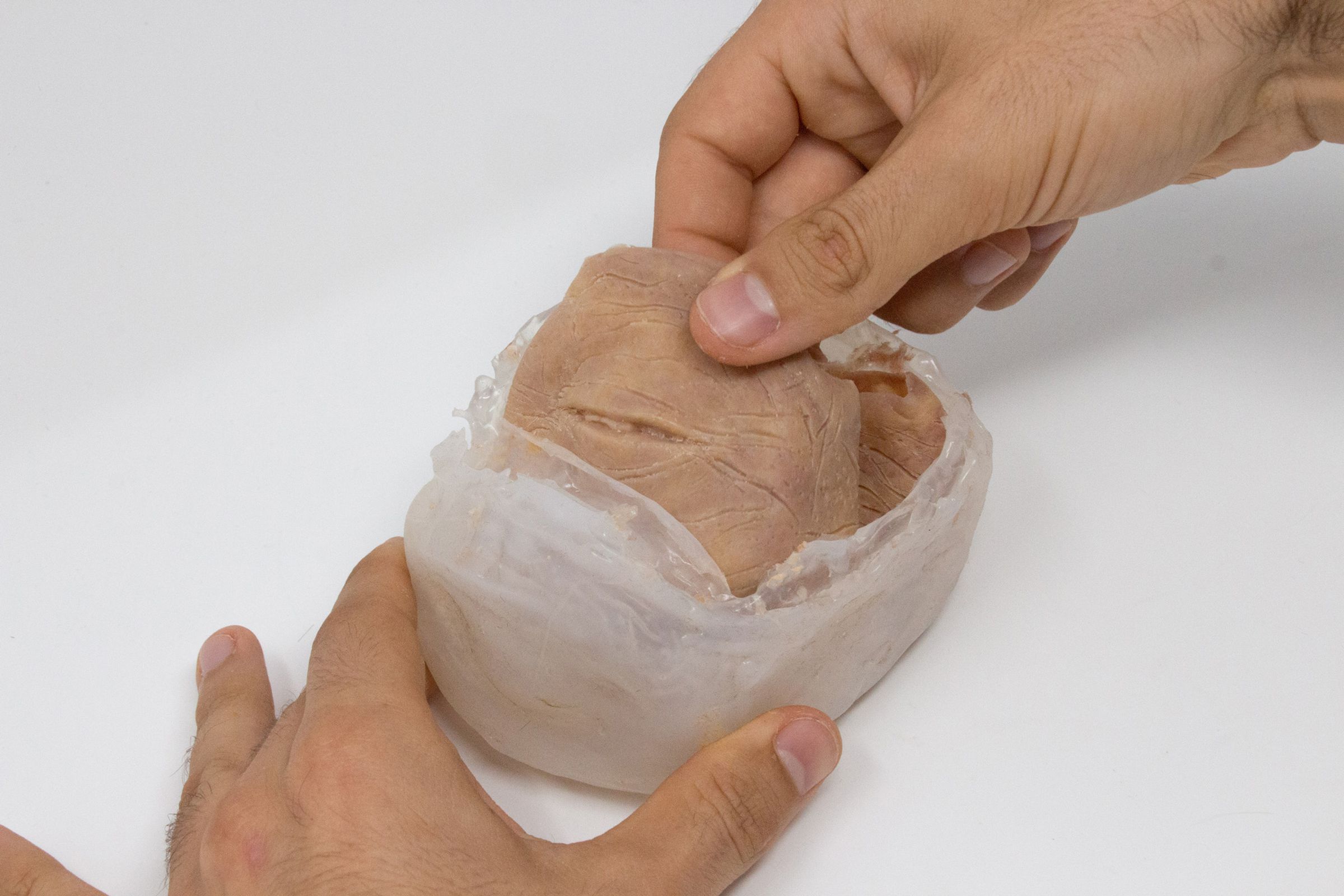 Two hands, one braced at the bottom of a container, and the other pulling out a chunk of synthetic flesh with eyelids. The eyeball has not been inserted yet. 