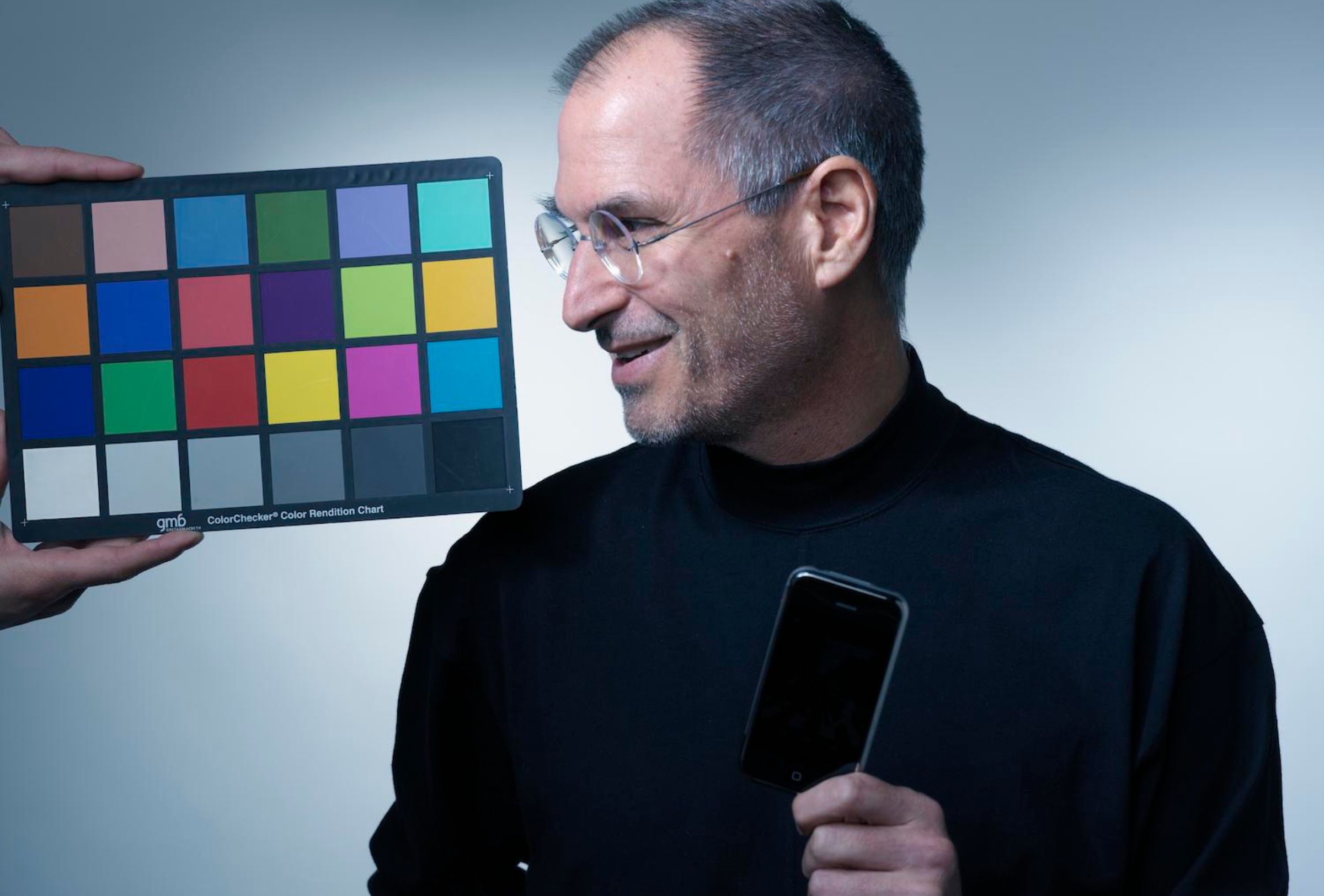 A photograph of Steve Jobs holding the first-generation iPhone in 2007. Someone is holding a color-correction card besides Jobs.