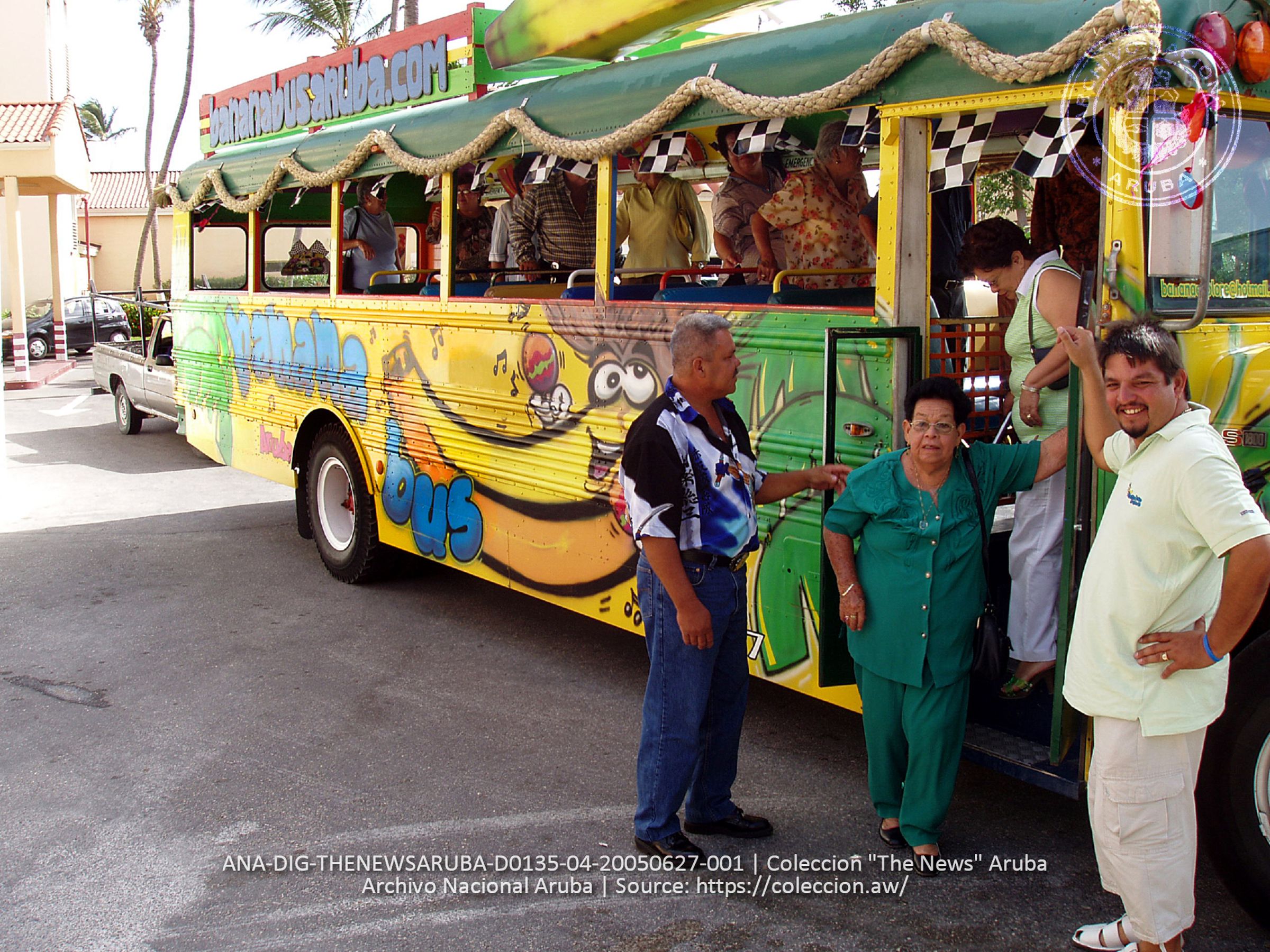 A picture of two volunteers standing by a colorful bus that reads “Banana Bus” on the side and has a banana holding a maraca. Two elderly women are stepping off of the bus.