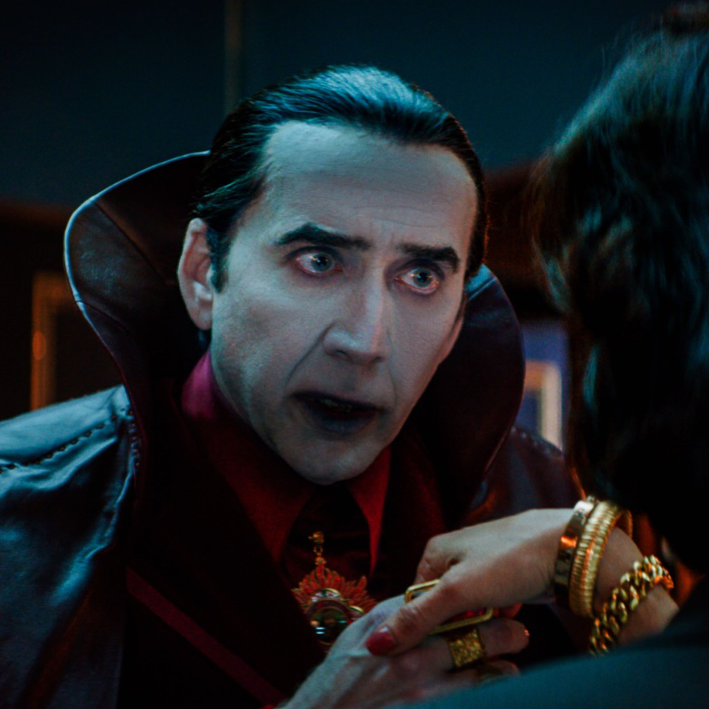 A vampire in a high-collared cape pulling a woman’s hand up to his mouth to kiss it.