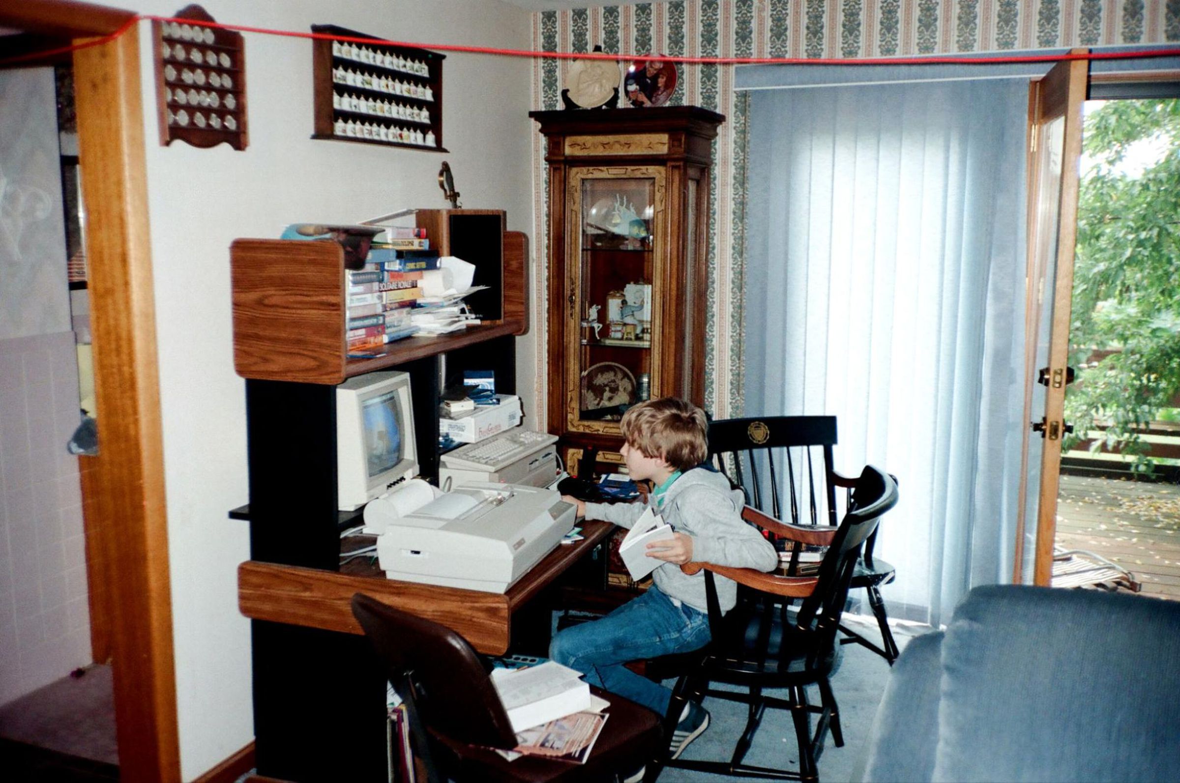 vintage image of living room with wallpaper, blue carpet, and open door to back yard. with blue cloth sofa and classic desk with computer, classic crt monitor, and printer with paper roll and kid using the computer.
