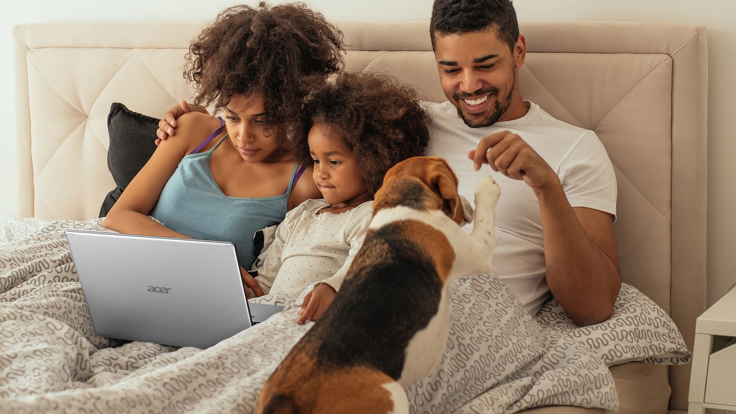 Two parents and a young child watch content on the Acer Aspire 5 in a bed. A dog paws one of the members.