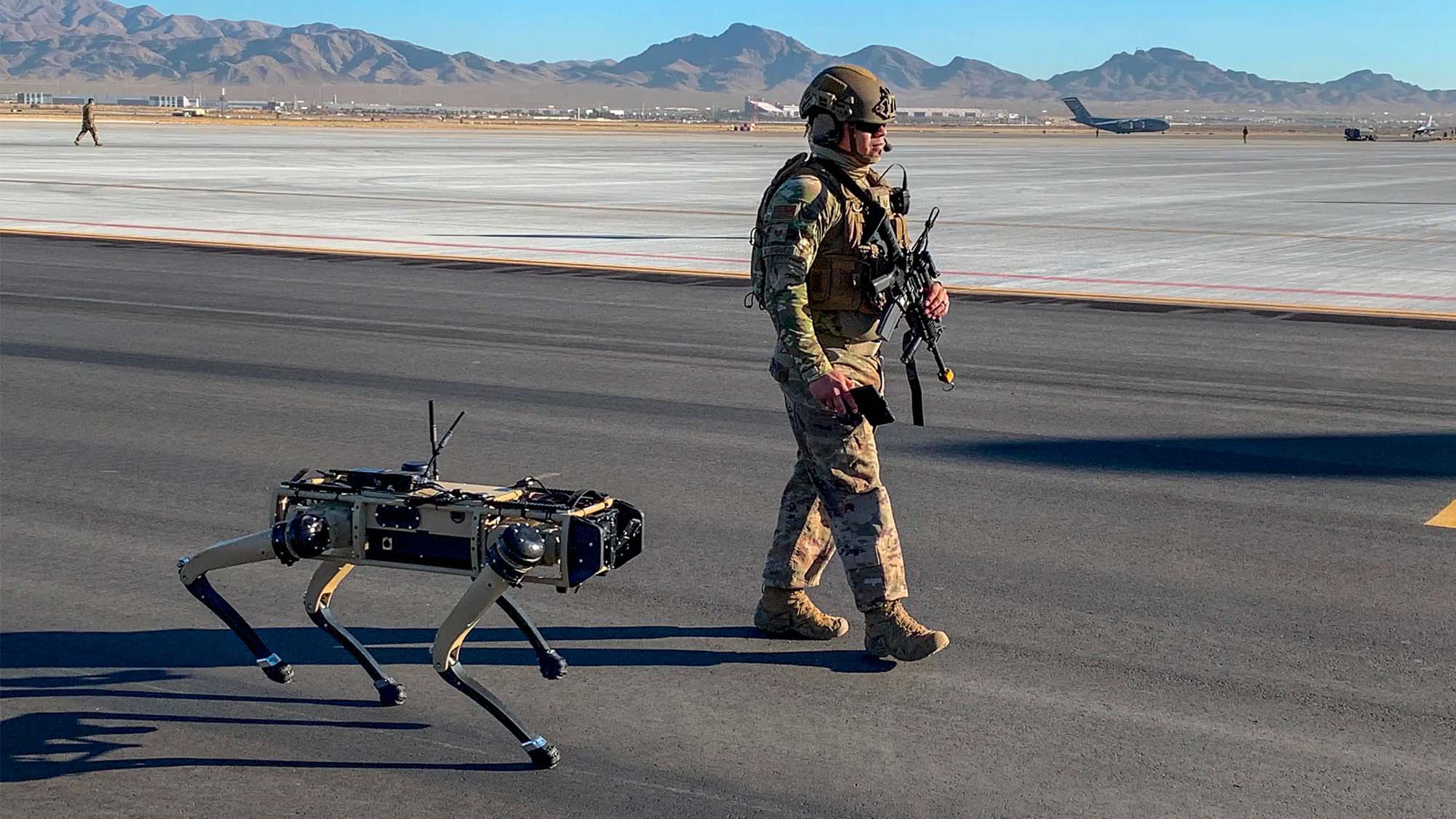 A photograph of a quadrupedal robot the size of a dog walking alongside an armed member of the Air Force on a US military base. 