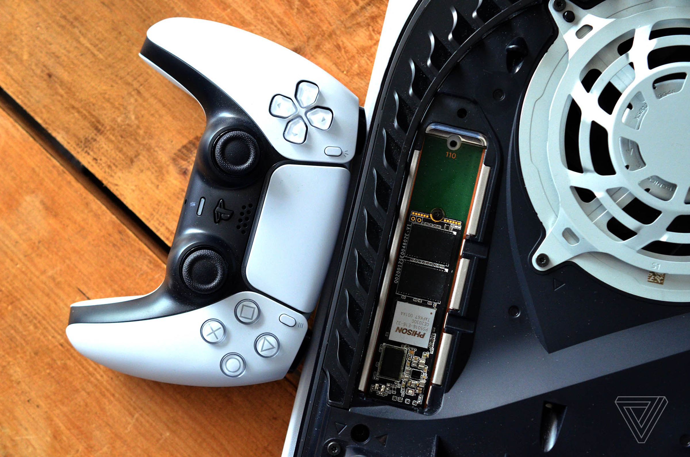 A close-up of a partially opened PlayStation 5 console, showing a user-installed SSD, with a DualSense controller sitting beside it.