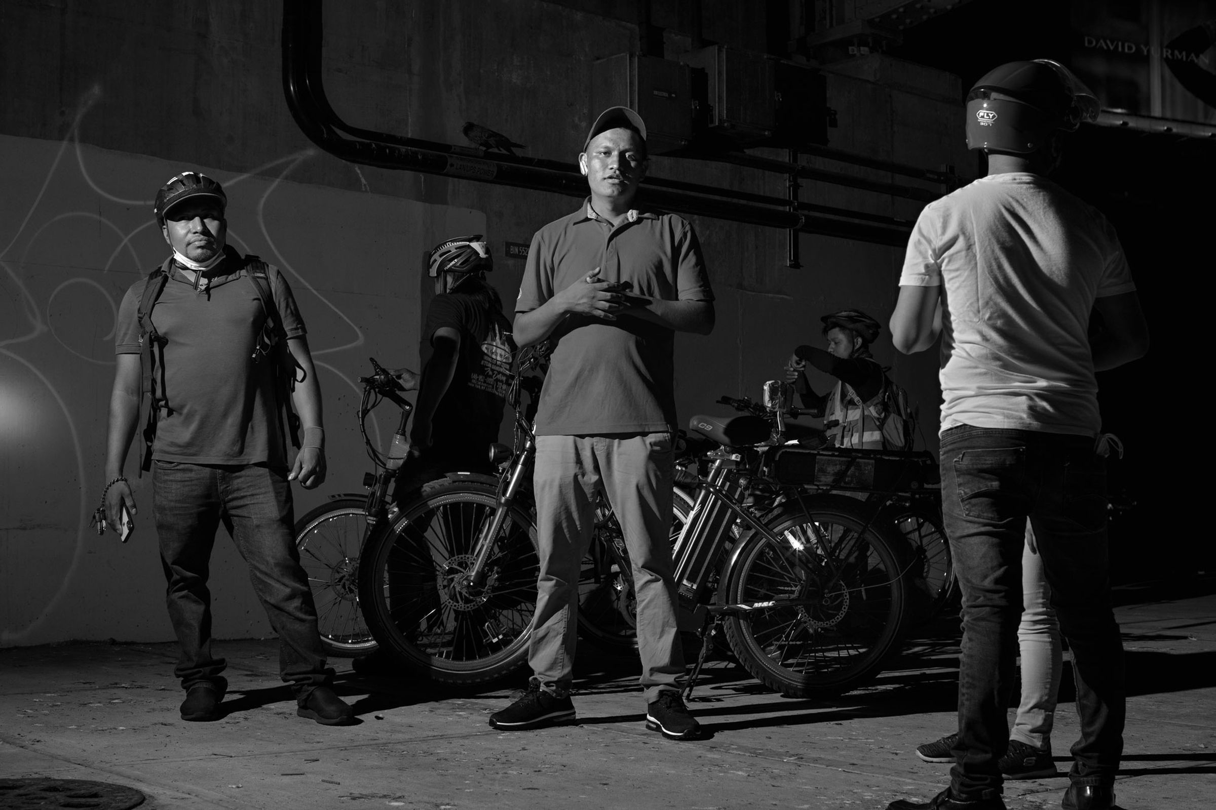 A black-and-white image of three delivery workers dressed in casual clothing, their bikes visible in the background.