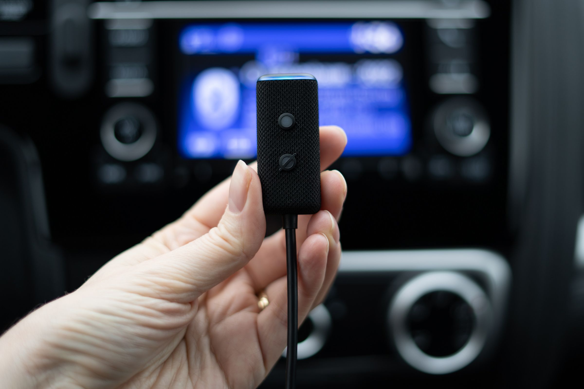 A close-up of a hand holding an Echo Auto, a small black device about 1 inch wide and 2 inches long. It has a small button and an indicator LED that leads to a cable. The photo was taken inside the car, with the center dash/infotainment area out of focus in the background.