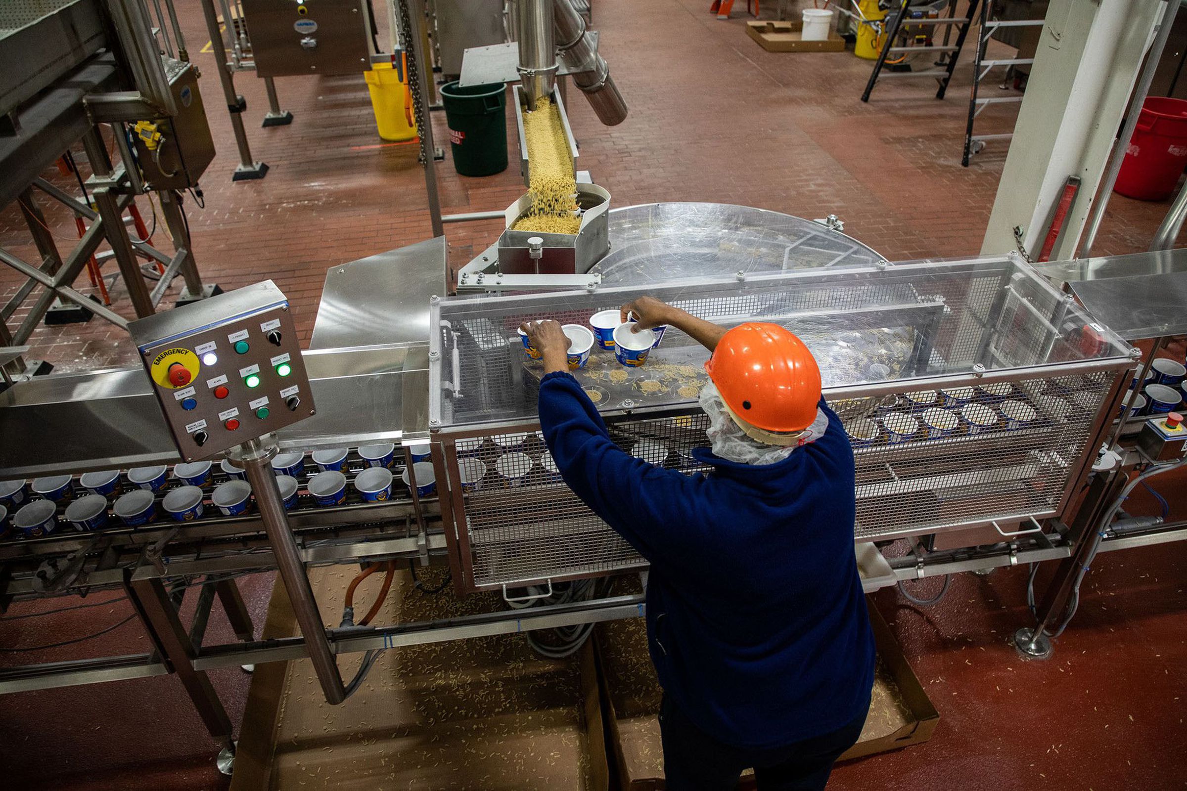 An employee wearing a hard hat and hair net is seen handling cup packaging on a macaroni production line.