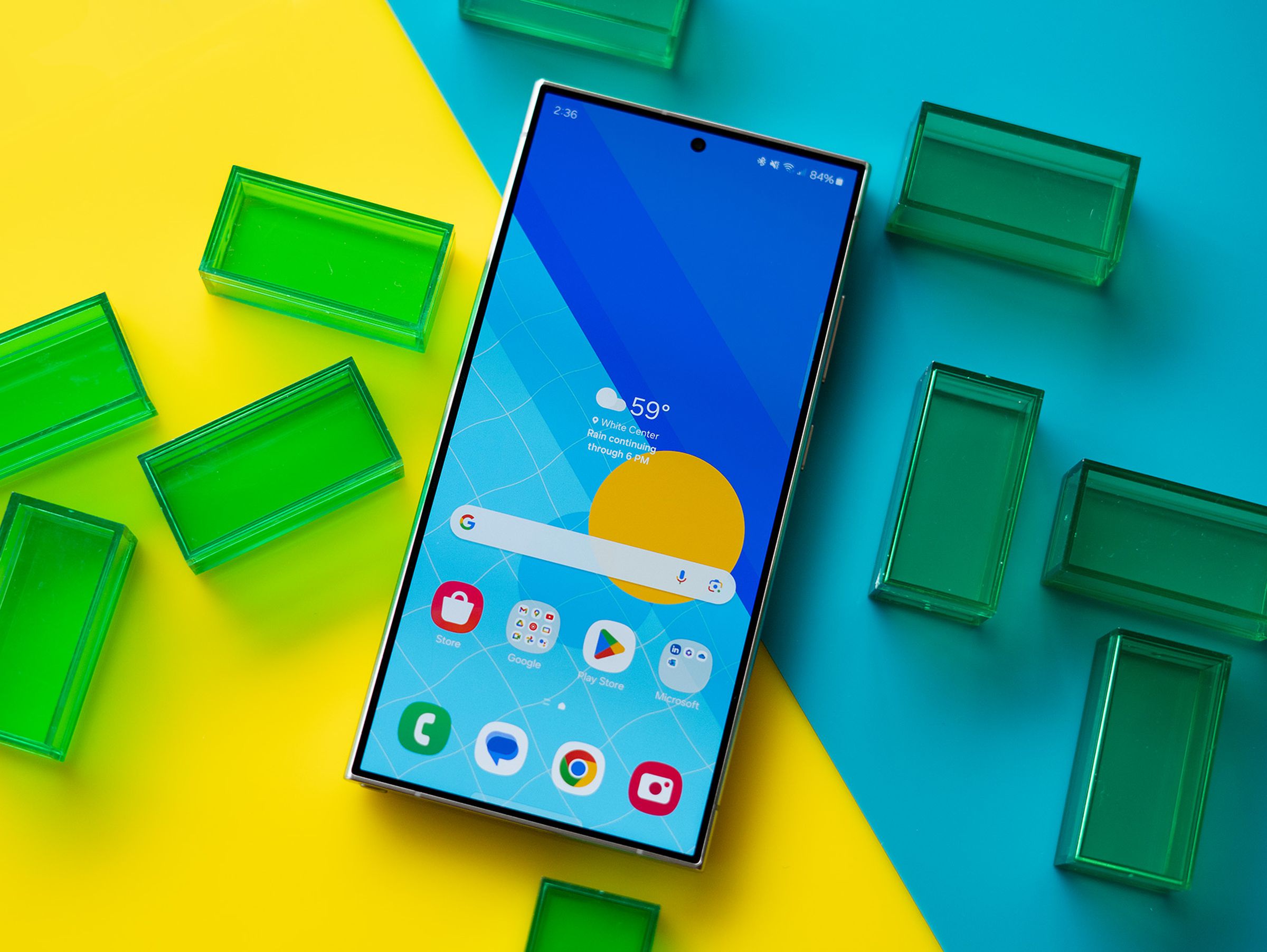 Samsung Galaxy S24 Ultra showing a blue and yellow homescreen, on a blue and yellow background with green translucent rectangles.