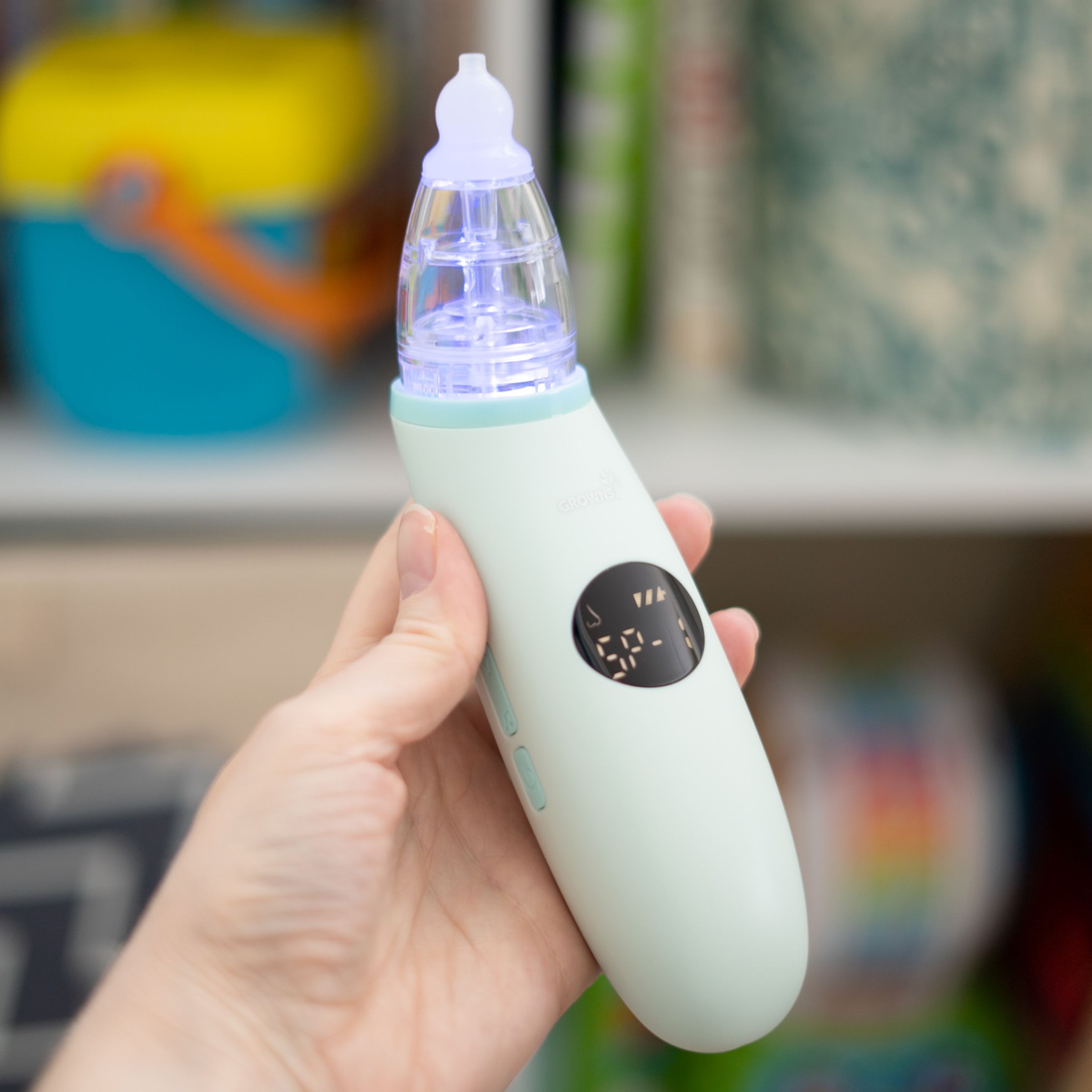 Hand holding a nasal aspirator in front of a colorful background of childrens’ toys and books.