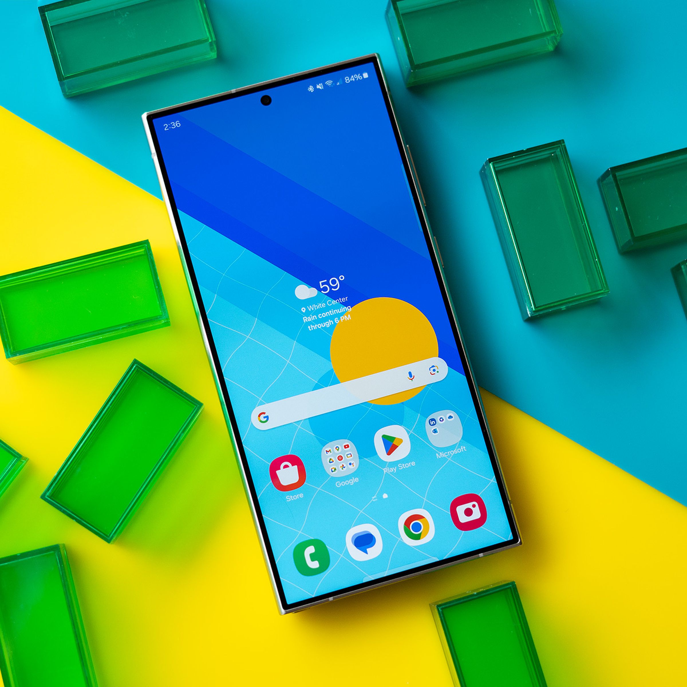 Samsung Galaxy S24 Ultra showing a blue and yellow homescreen, on a blue and yellow background with green translucent rectangles.