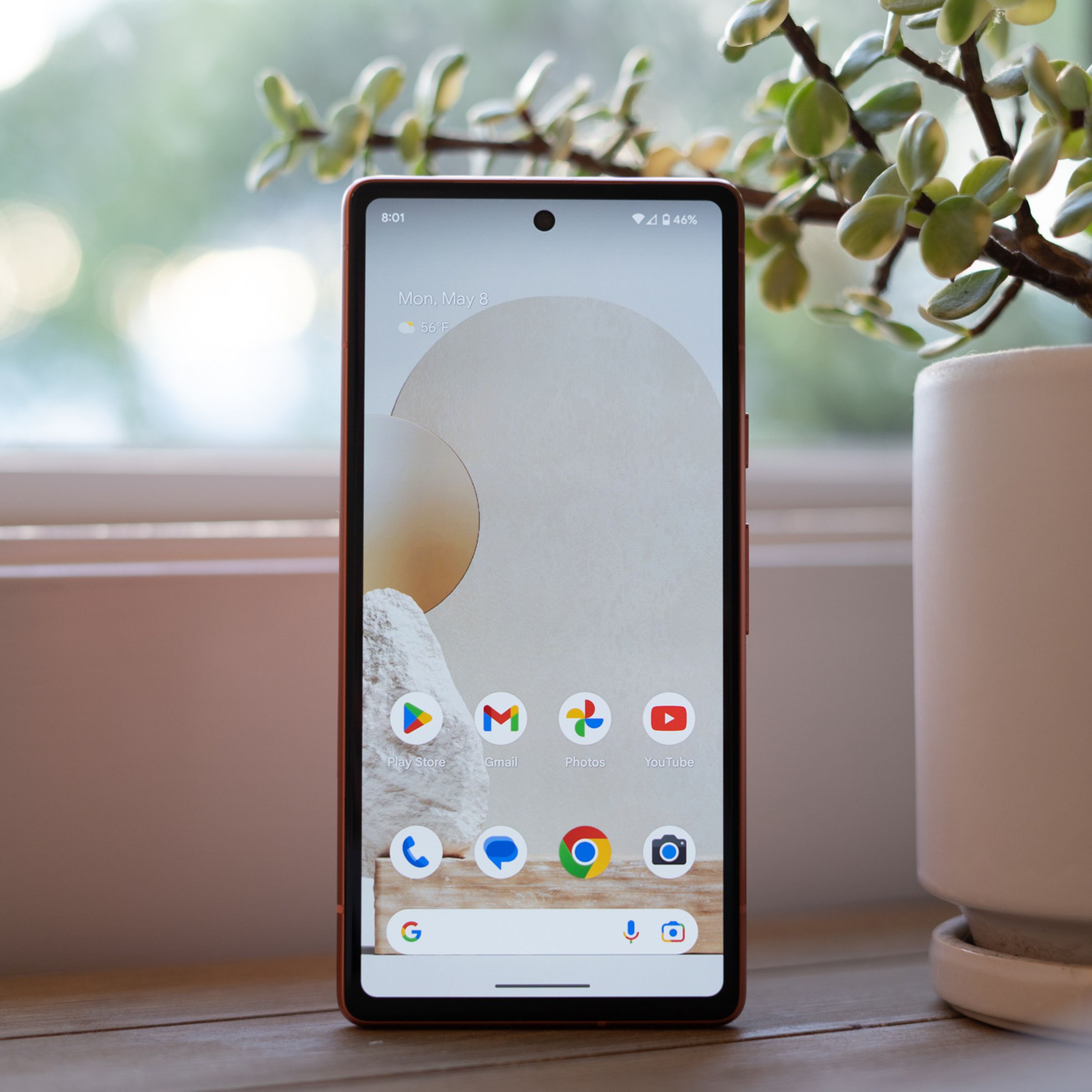 Google Pixel 7A standing upright on a table and displaying the home screen.