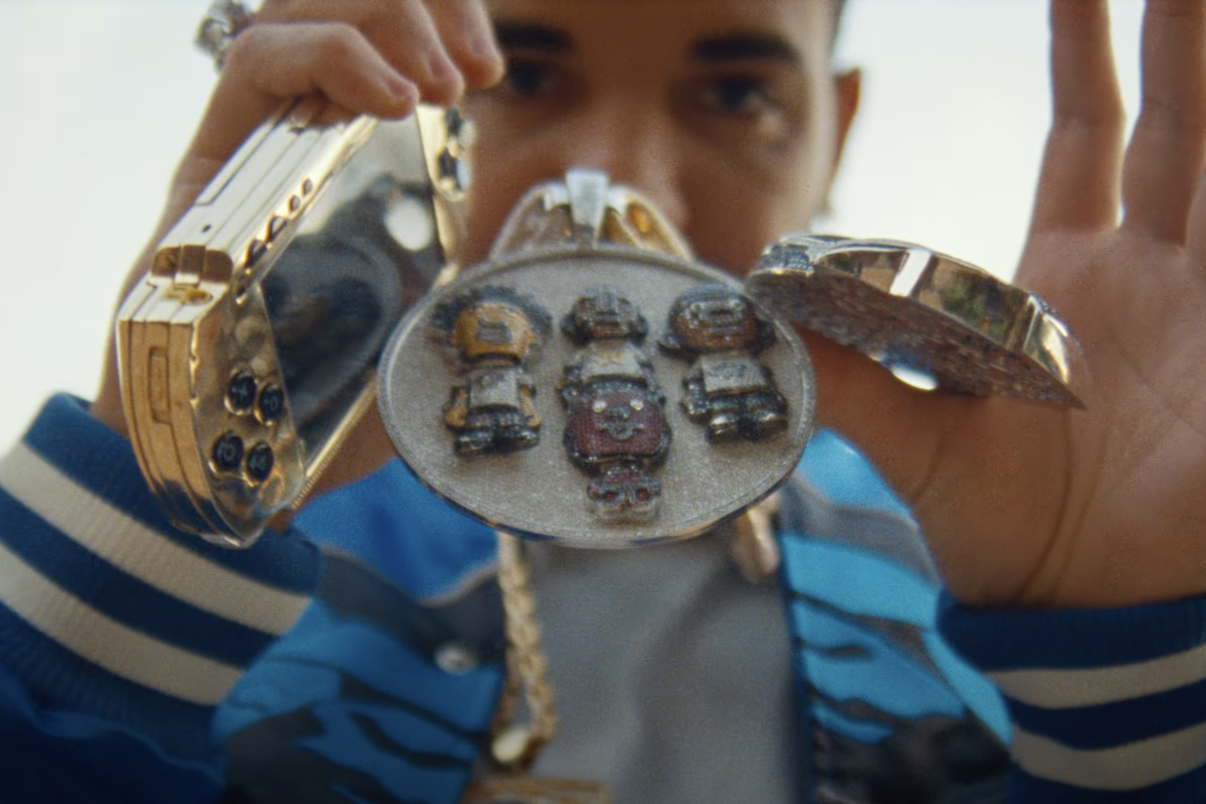 A close-up of a gold PSP and Pharrell Williams’ chains / medallions in the hands of Drake, who is holding it all right in front of his face.