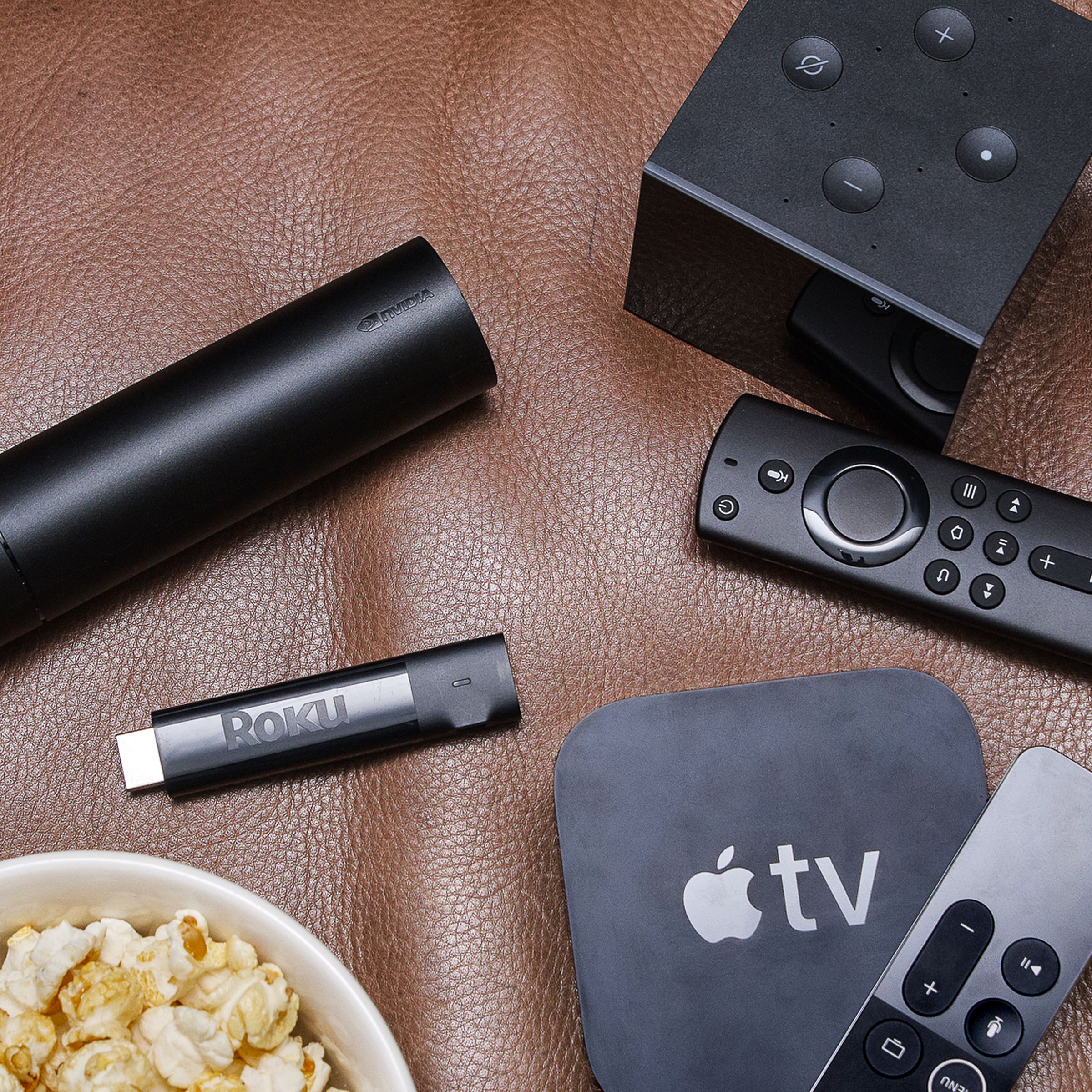 The Apple TV 4K, Amazon Fire Cube, Roku Streaming Stick, and Nvidia Shield streaming devices and remotes laid out on a brown leather couch next to a bowl of popcorn.
