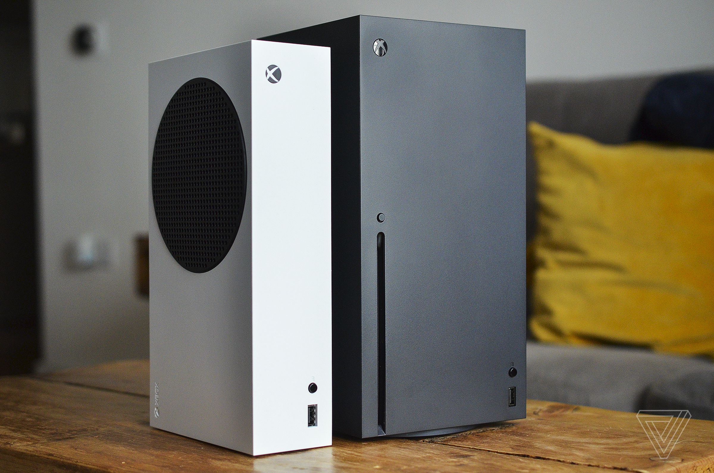 Die weiße Xbox Series S von Microsoft steht neben der größeren, schwarzen Xbox Series X auf einem hölzernen Couchtisch im Wohnzimmer