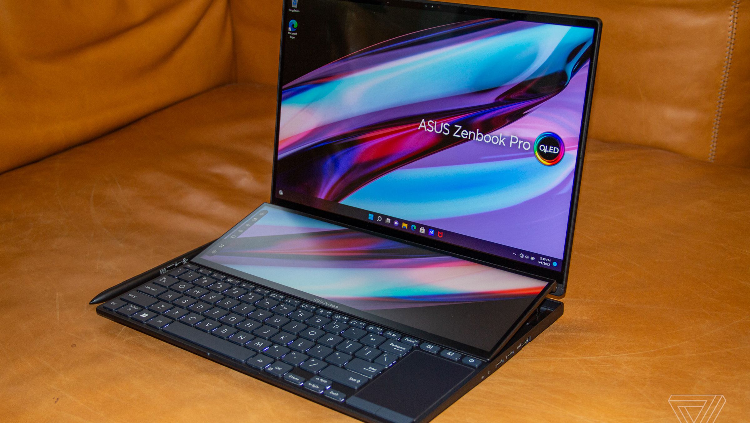 The Asus Zenbook Pro 14 Duo angled to the left side on a brown couch. Both screens display a multicolor background with the Asus Zenbook Pro OLED logo.