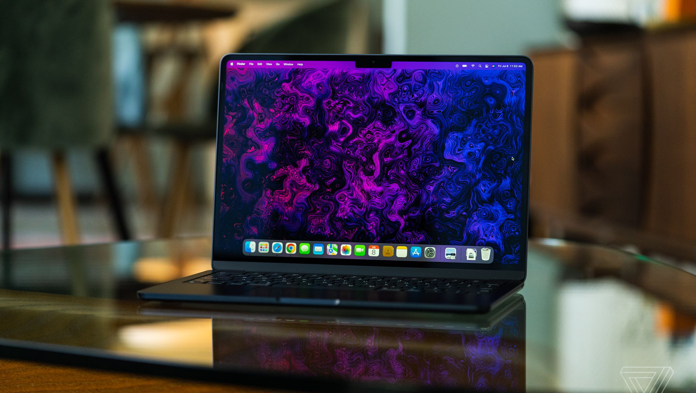 The M2 MacBook Air is opened, facing the camera. Its display is on, showcasing a psychedelic purple and black wallpaper created by The Verge’s art and illustration team.