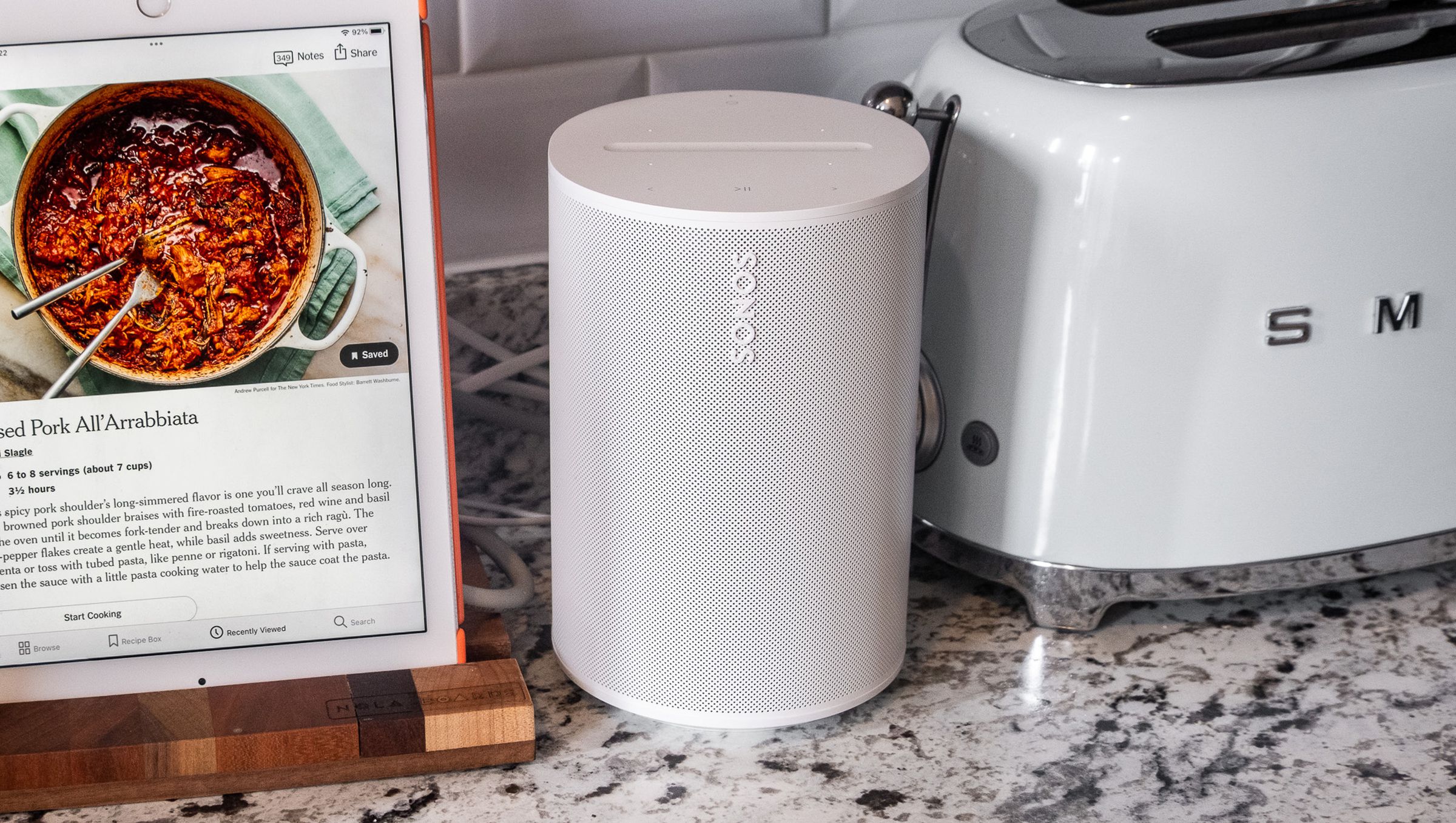 A photo of the Sonos Era 100 speaker in a kitchen setting beside an iPad and toaster.
