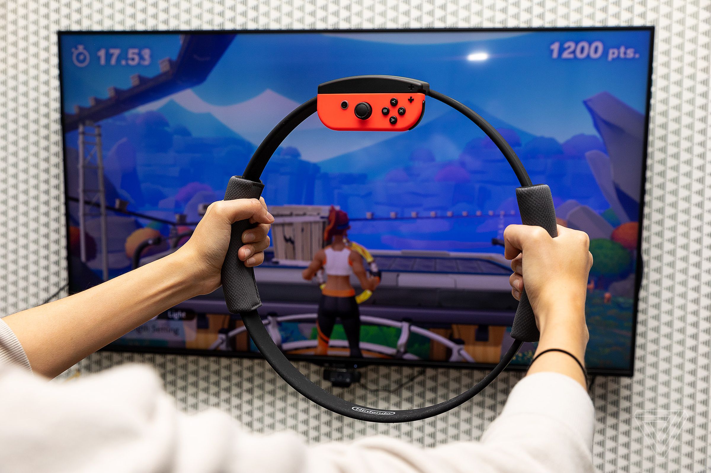 A person holding a Ring Fit Adventure pilates ring controller in front of a wall-mounted TV playing the game.
