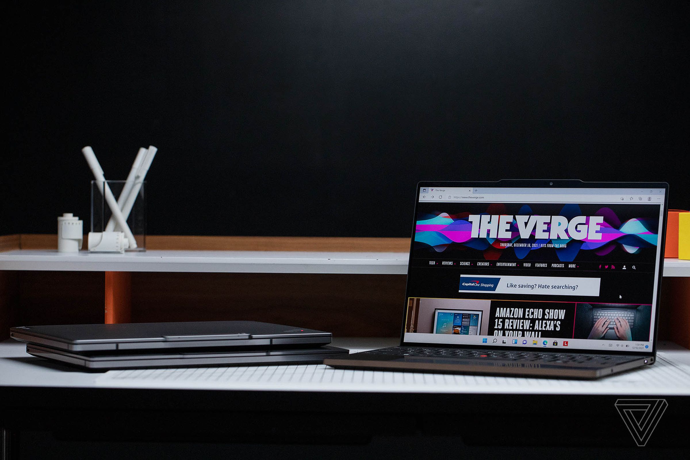 The ThinkPad Z13 open on a table with a black background displaying The Verge homepage. To its left are two more ThinkPad Z13 models stacked atop each other.