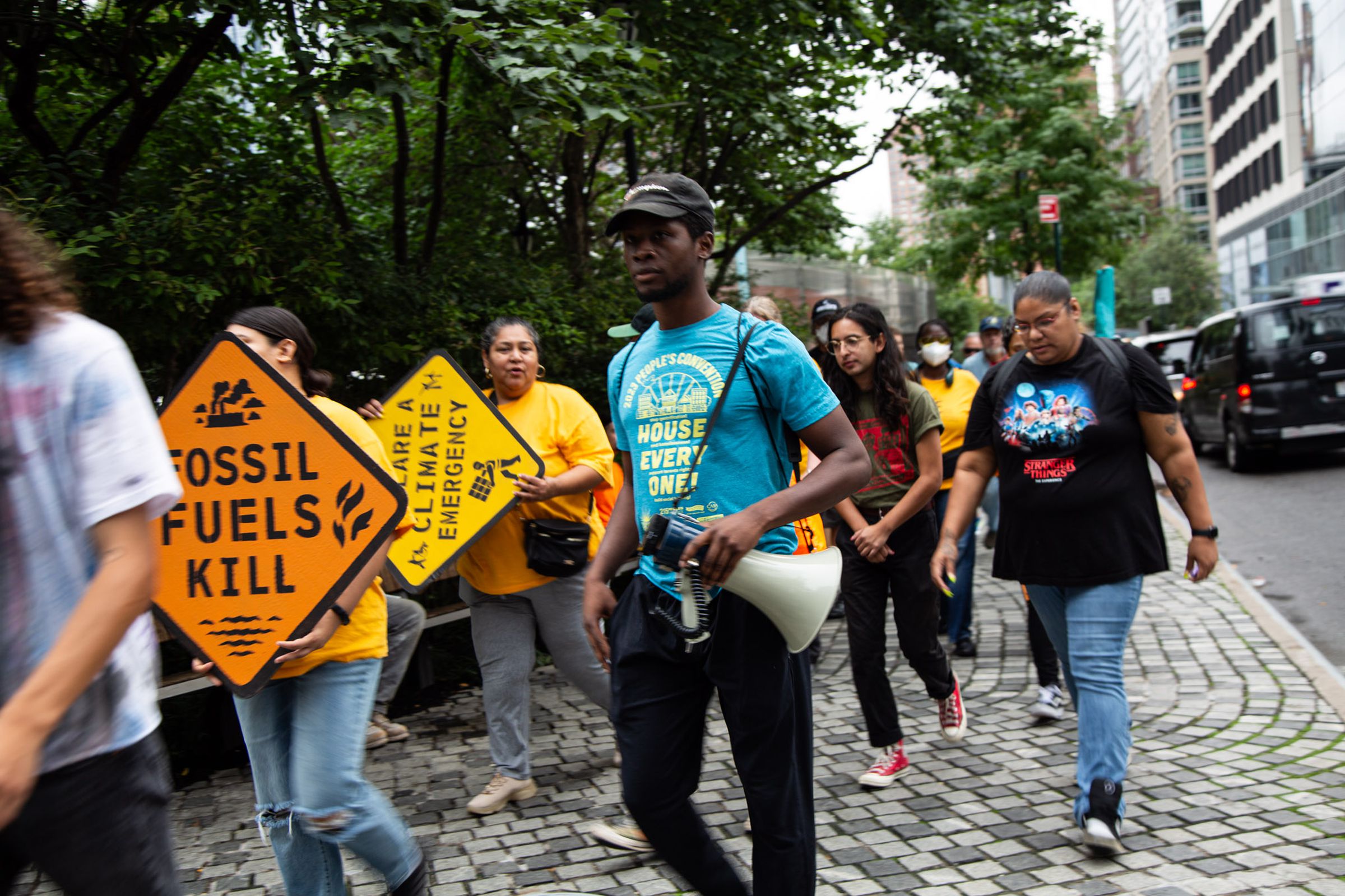 A group of people walk down the sidewalk. One carries a bullhorn, others carry signs that say “fossil fuels kill” and “ declare a climate emergency.”