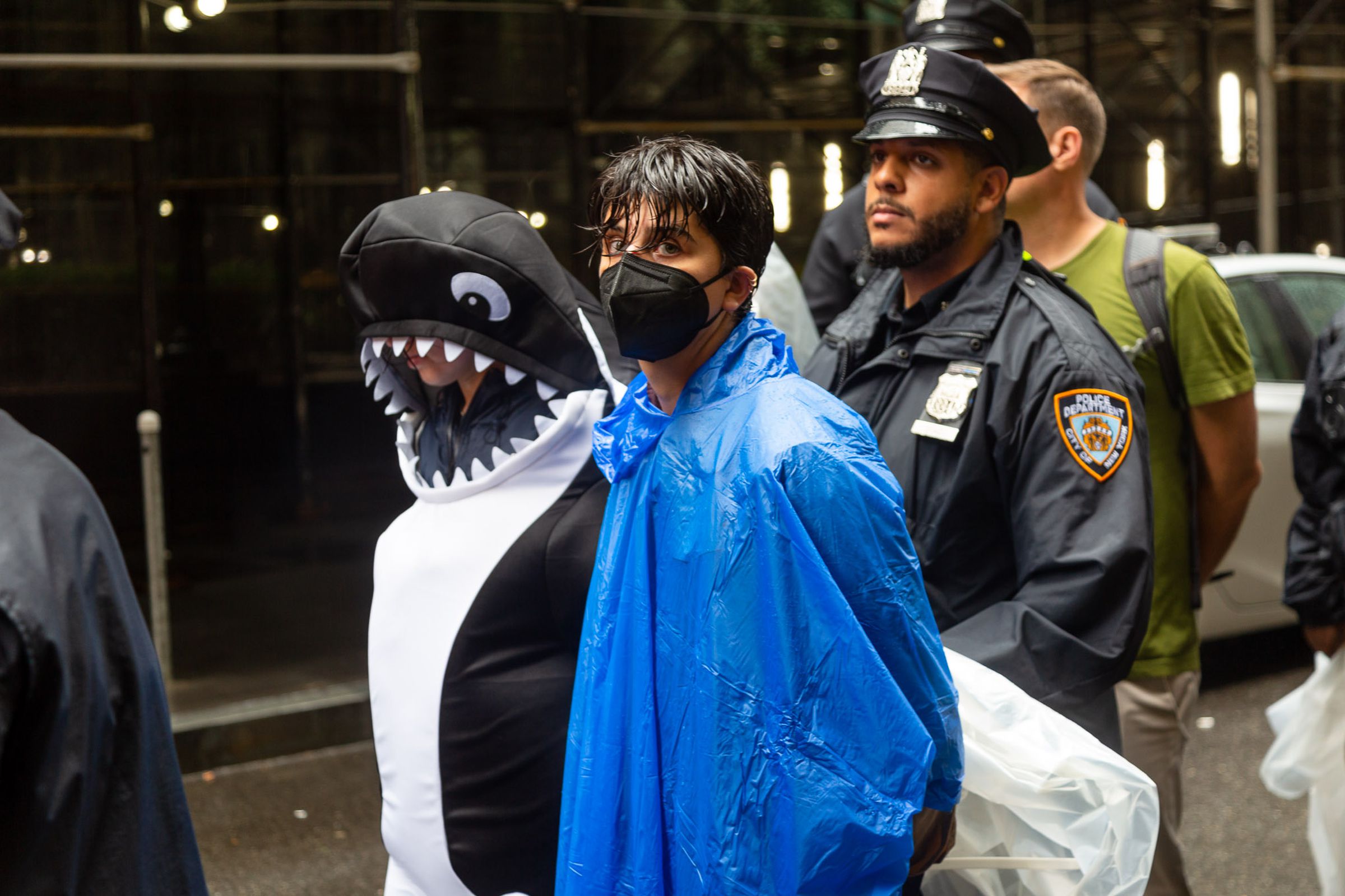 A protester dressed as an orca next to a protester wearing a blue poncho and mask. Both have their hands tied behind their back.